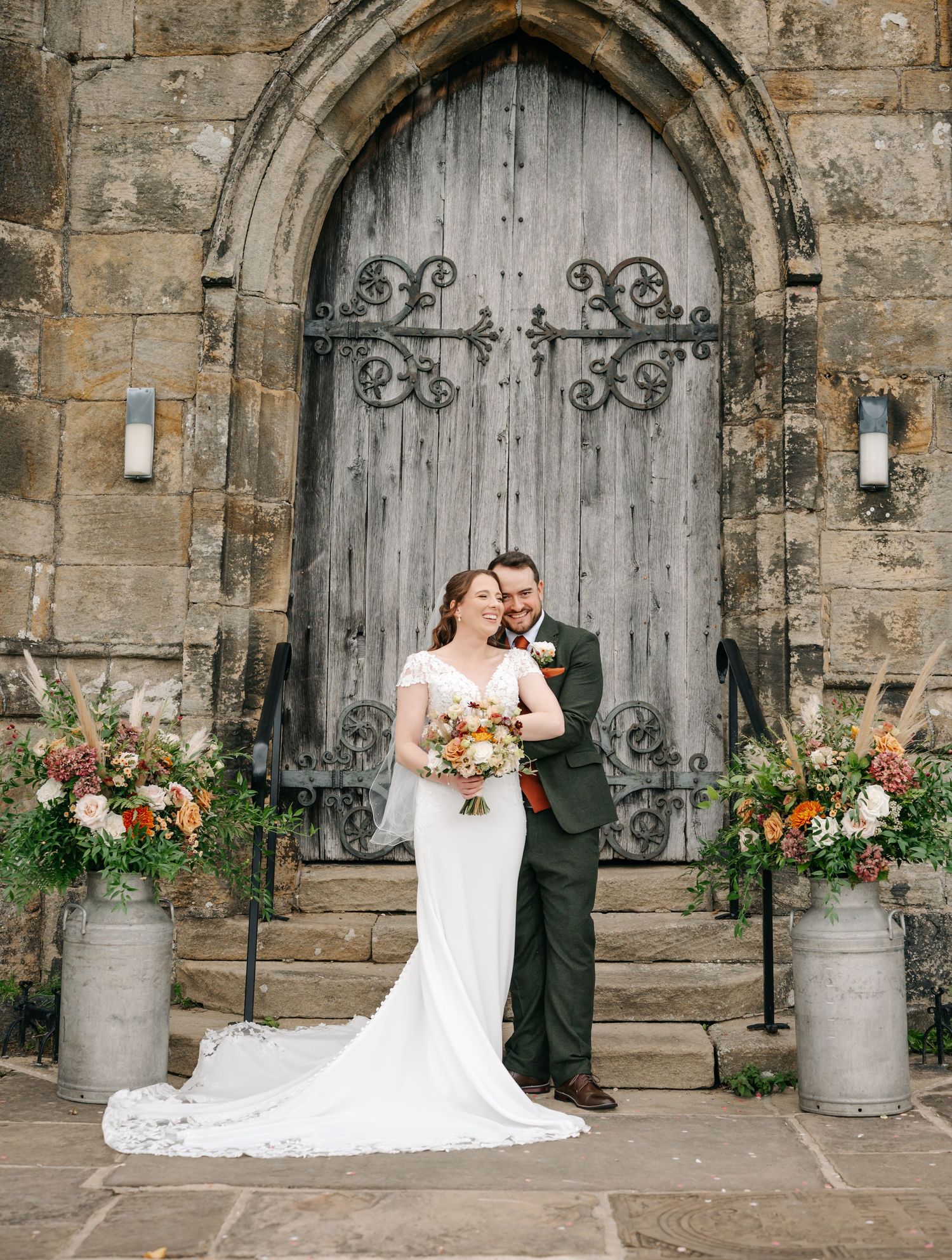 Bride in a white gown and groom in a green suit embrace on stone steps before a rustic arched wooden church door with floral arrangements.