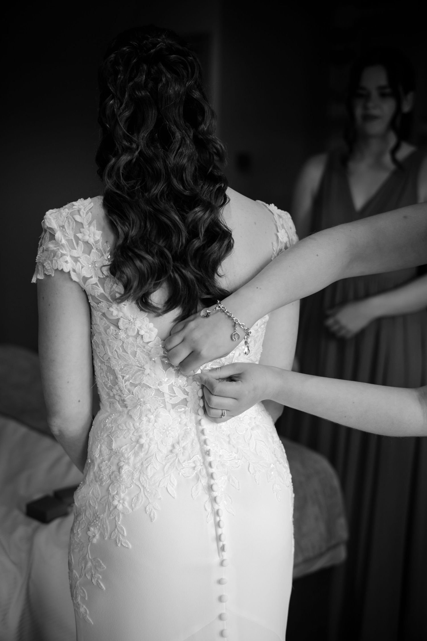 Black and white photo of bridesmaids fastening the buttons on the back of a lace wedding dress.