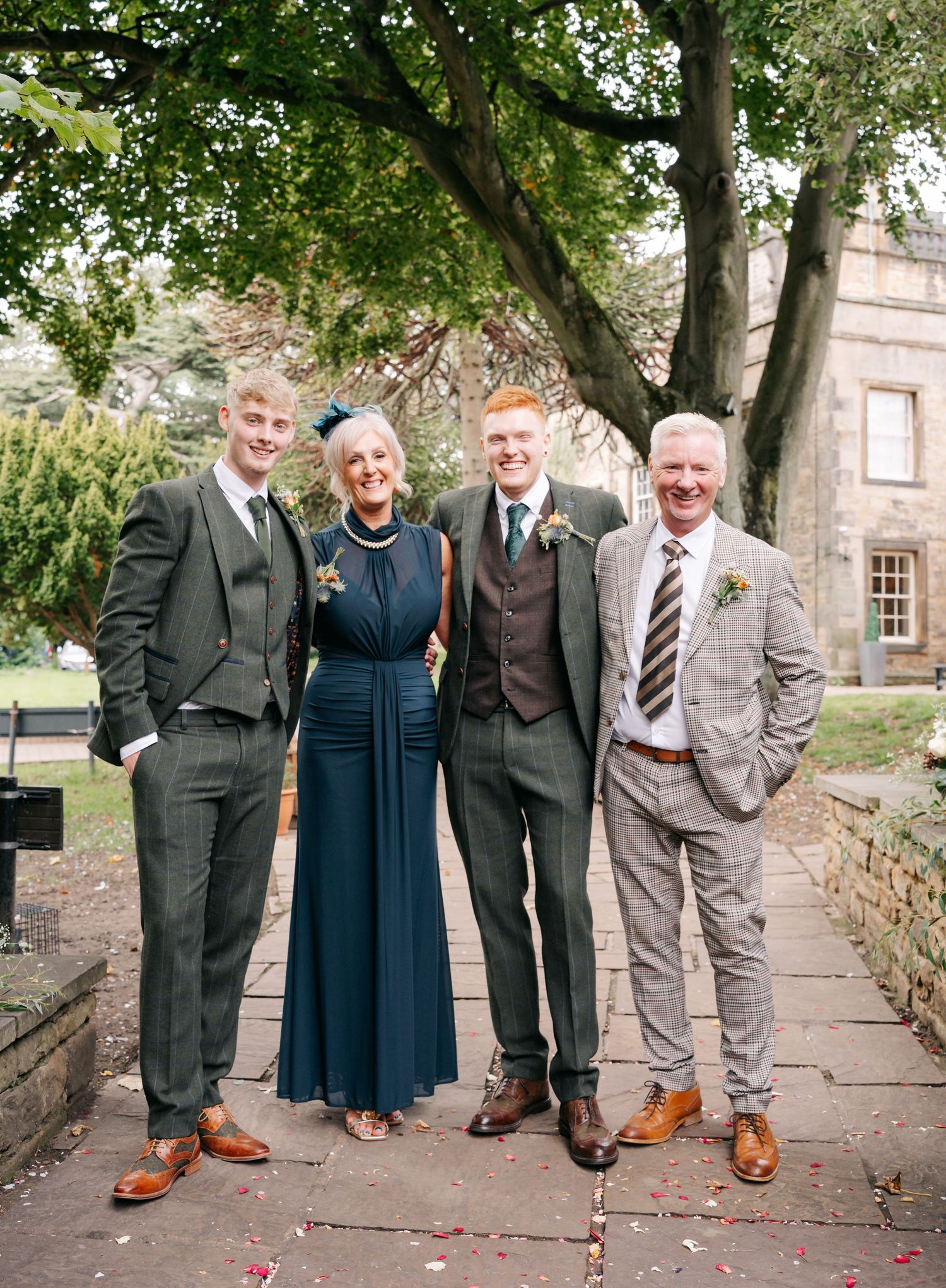 Group photo of wedding party in formal attire on stone pathway under trees.