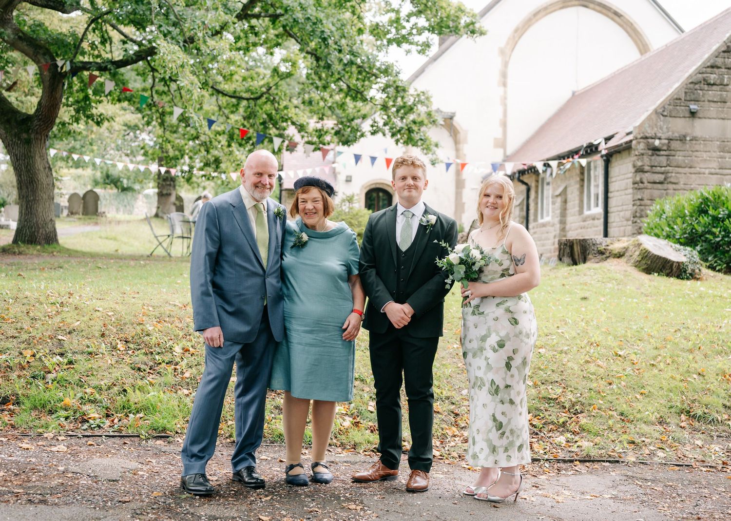 Four people in formal attire pose together on a lawn in front of a stone church archway.