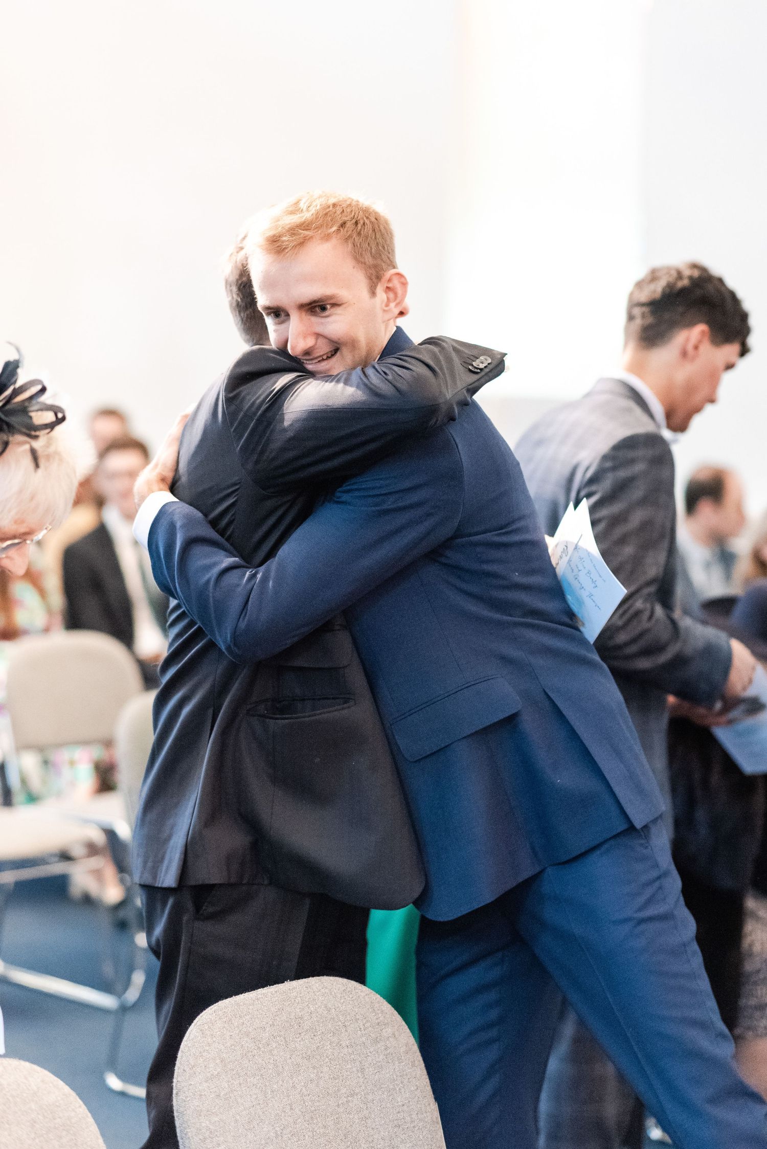 Wedding guests in navy suits share emotional embraces during a celebration.