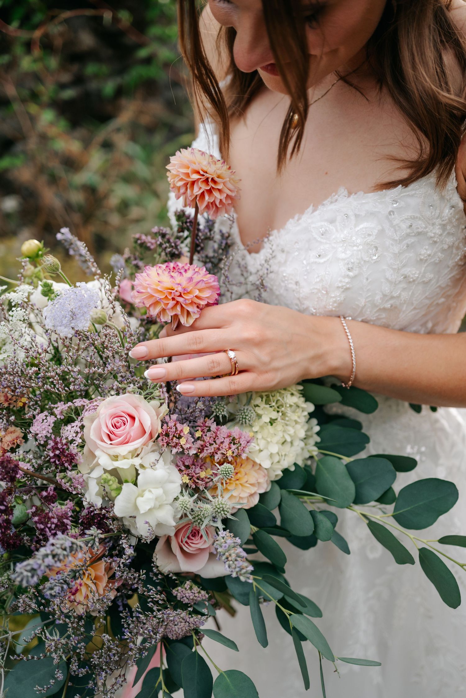 Bride in white lace gown holding lush wedding bouquet with pink roses, dahlias, and eucalyptus, wearing gold rings.
