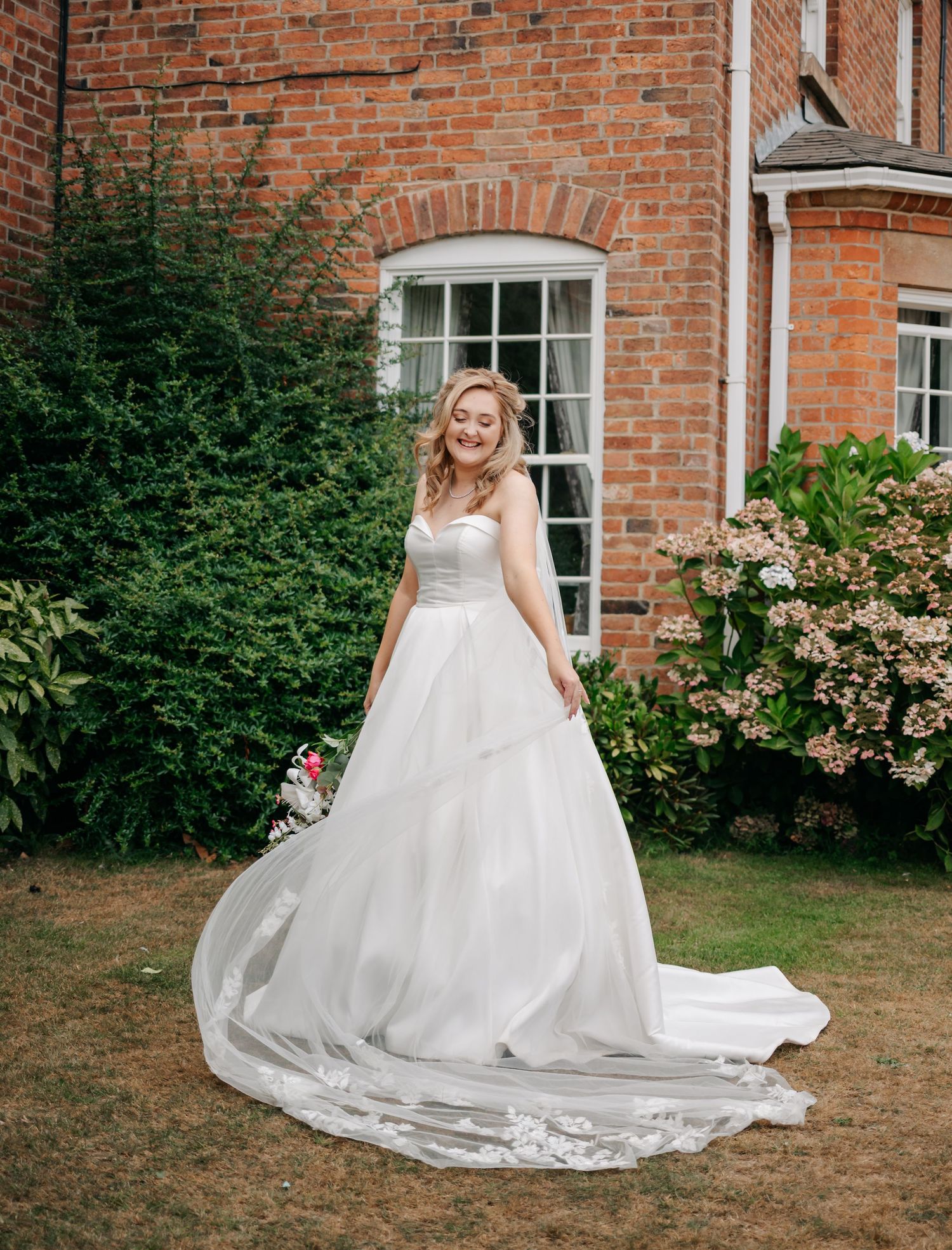 White wedding dress cascades across green lawn in front of classic brick building.
