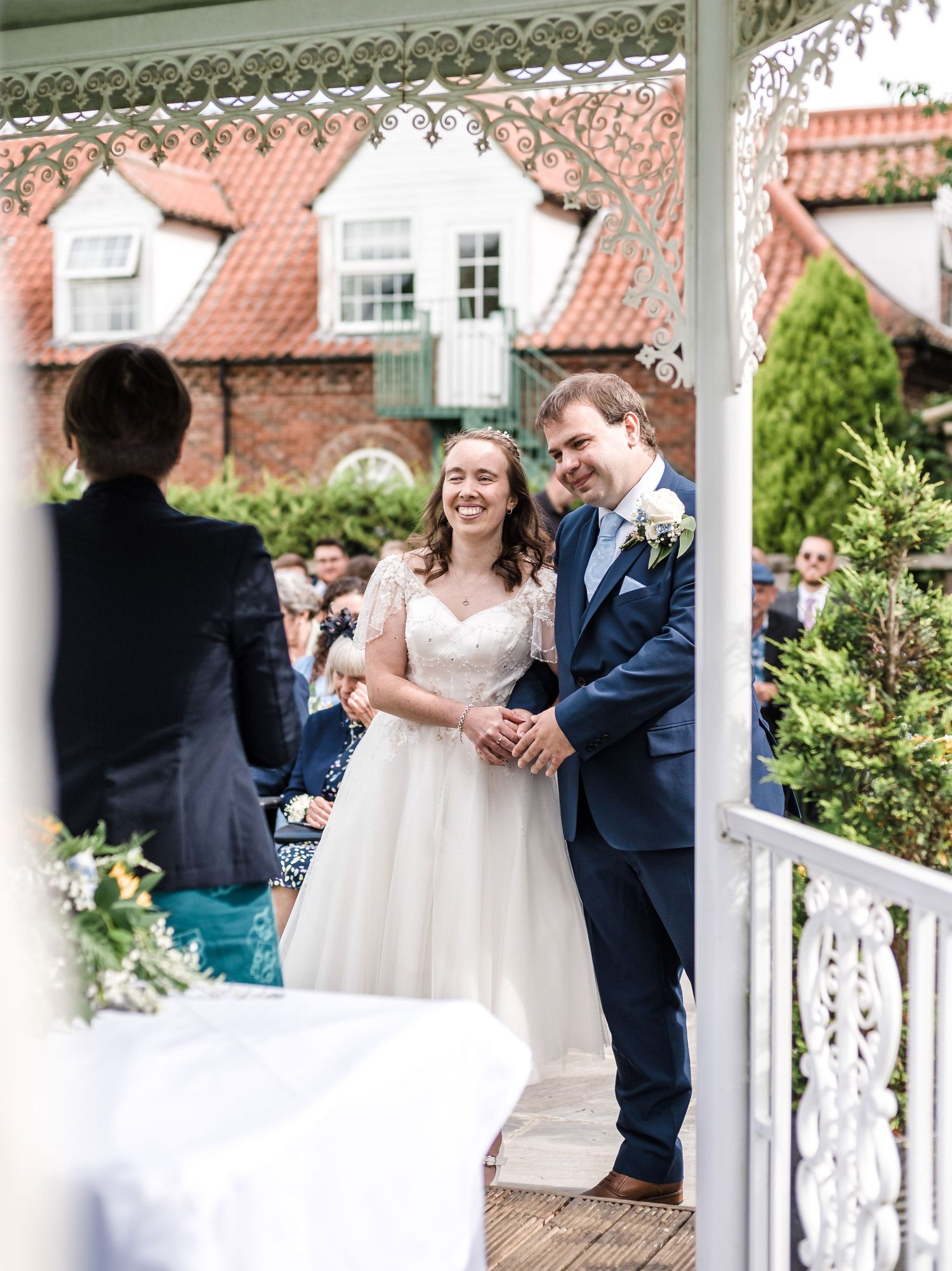 An outdoor wedding ceremony under a decorative gazebo with red-roofed buildings in the background.