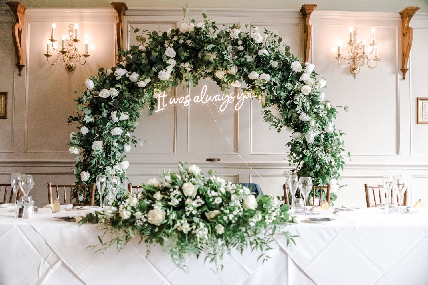 Stunning floral arch with white roses and greenery against an elegant white wall.