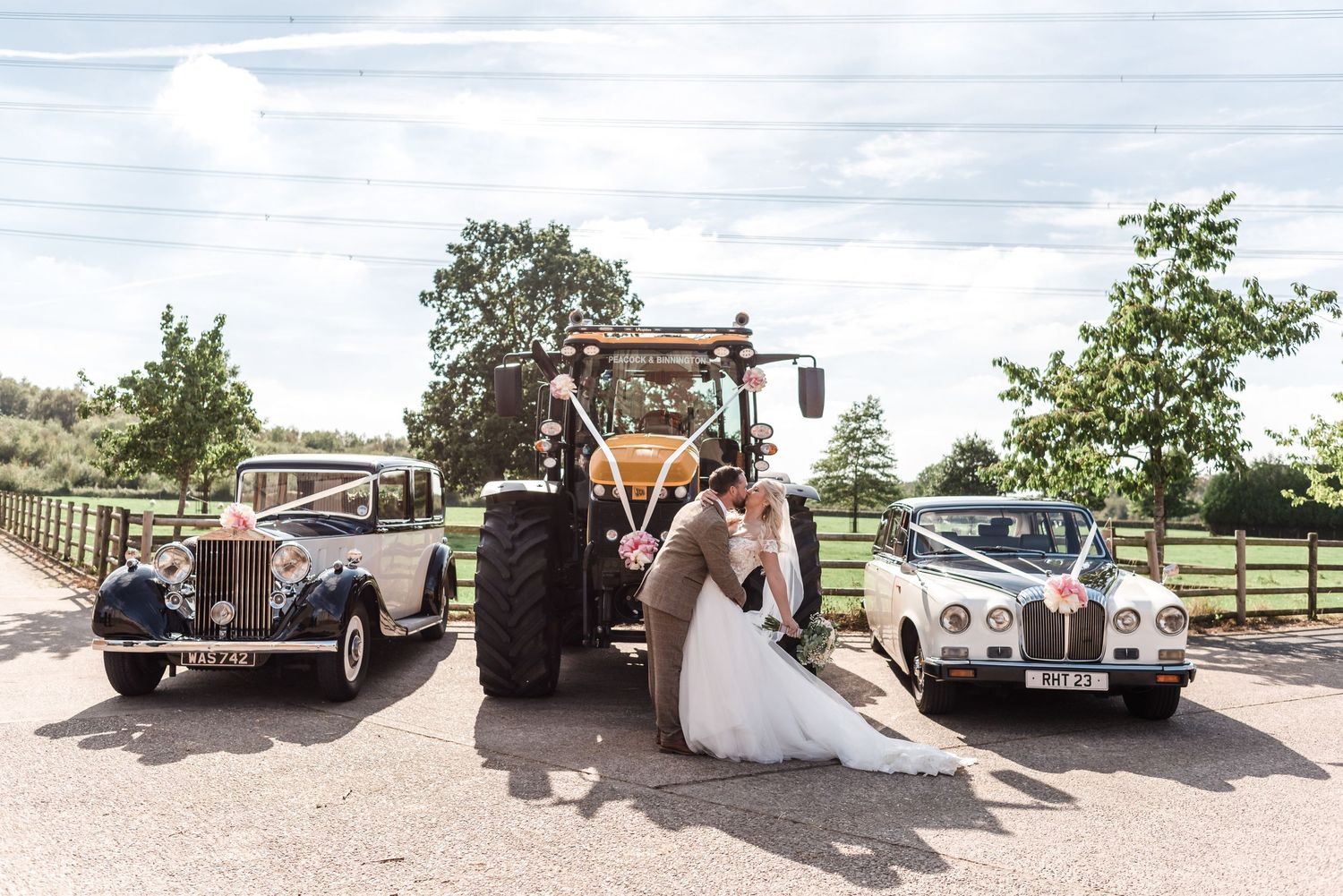 Wedding couple poses between vintage cars and tractor in rustic outdoor setting.