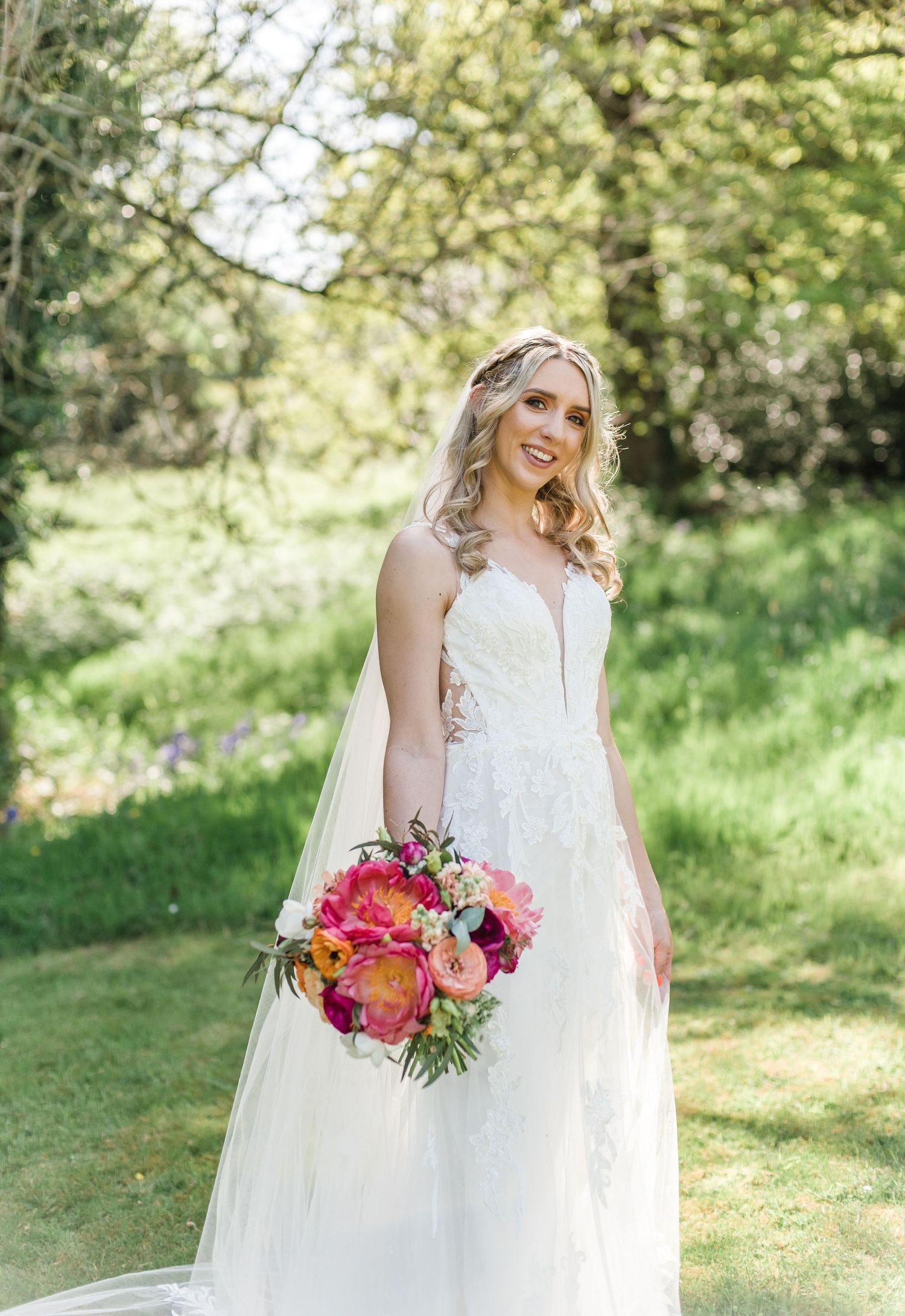 Elegant bride in white lace gown holds vibrant pink and coral bouquet in garden setting.