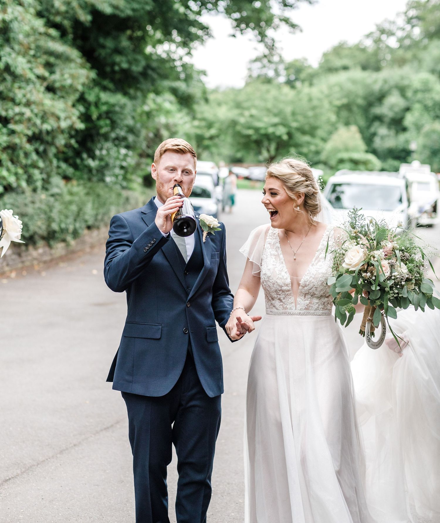 A couple in wedding attire walking down a tree-lined road sharing a joyful moment.