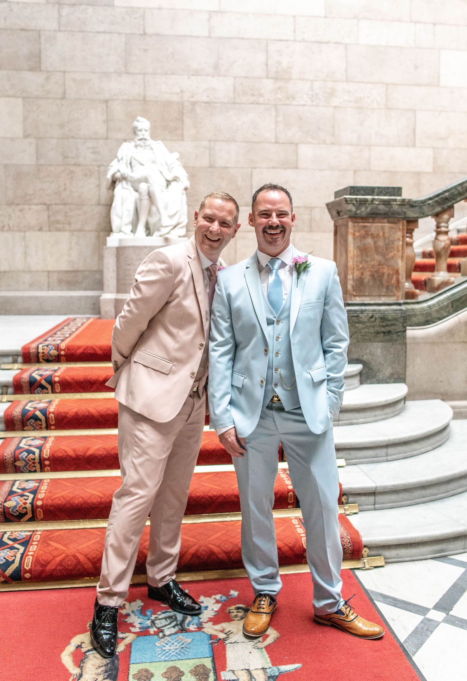 Wedding couple in matching light-colored suits pose on red carpeted steps in front of a stone building.