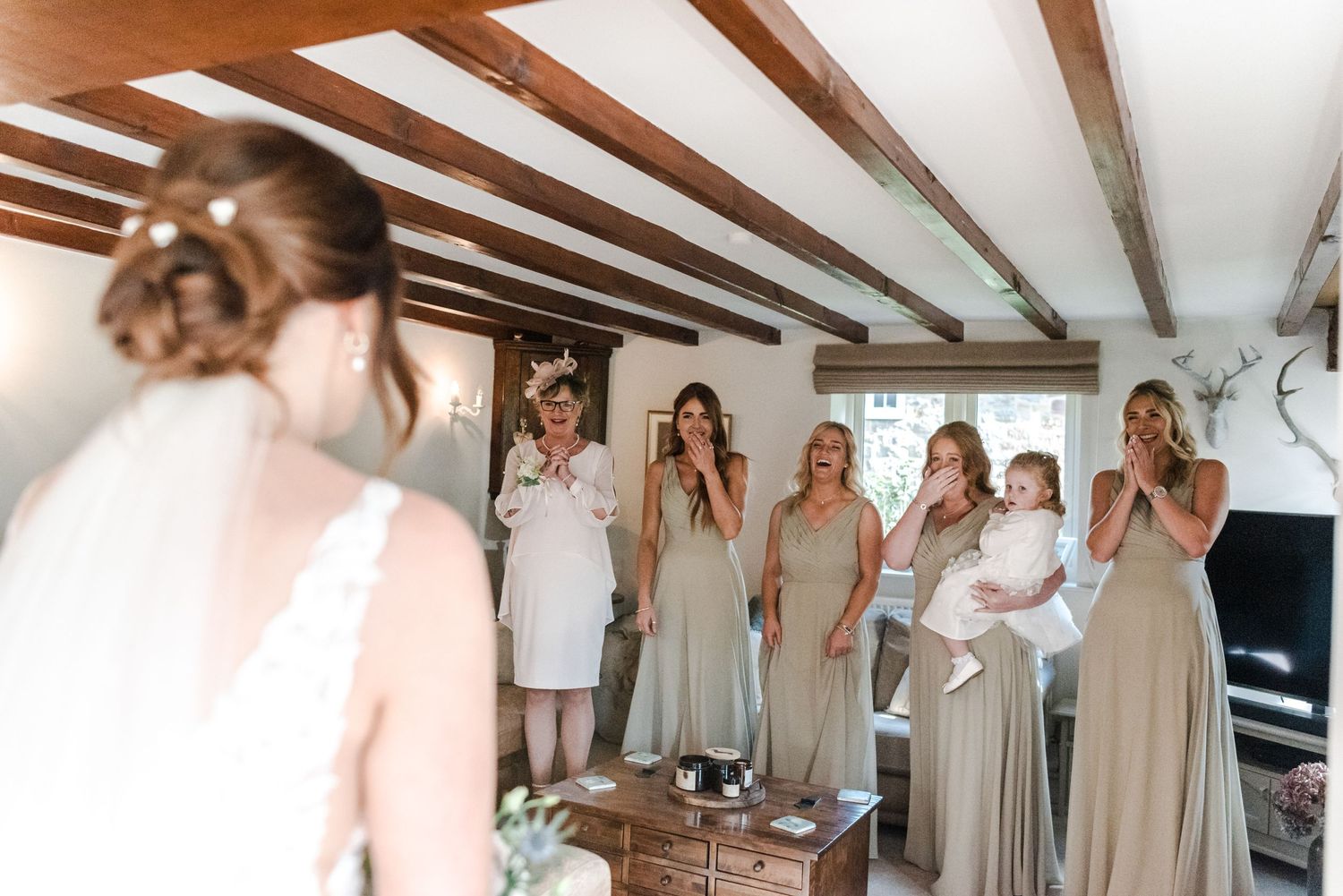 Bridesmaids in neutral dresses react with joy during wedding preparations.