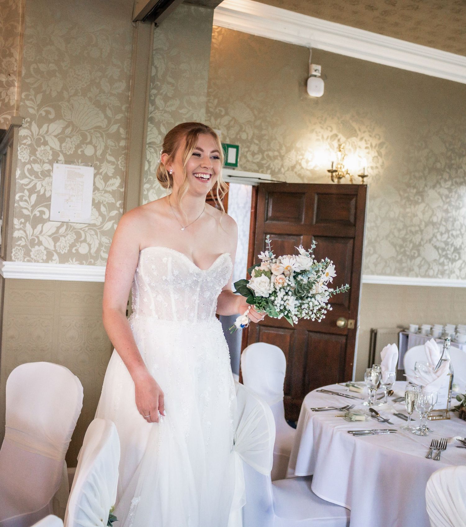 A person wearing a white strapless wedding gown smiles while standing in an elegant reception venue.