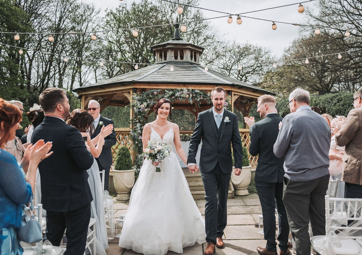 Wedding couple walks down the aisle while guests applaud during outdoor ceremony at a gazebo.