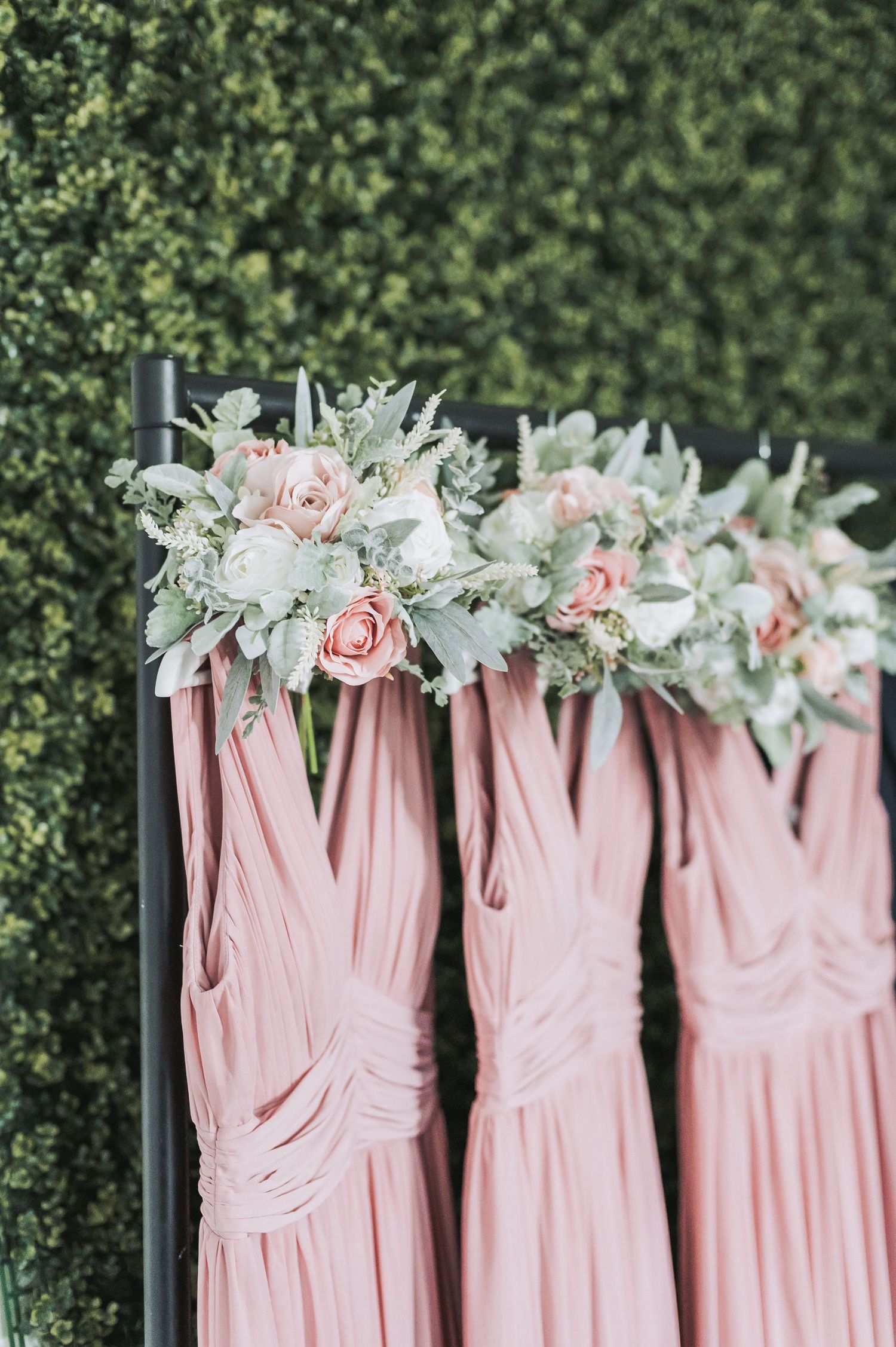 Pink bridesmaid dresses with delicate floral bouquets hang against a lush green hedge backdrop.