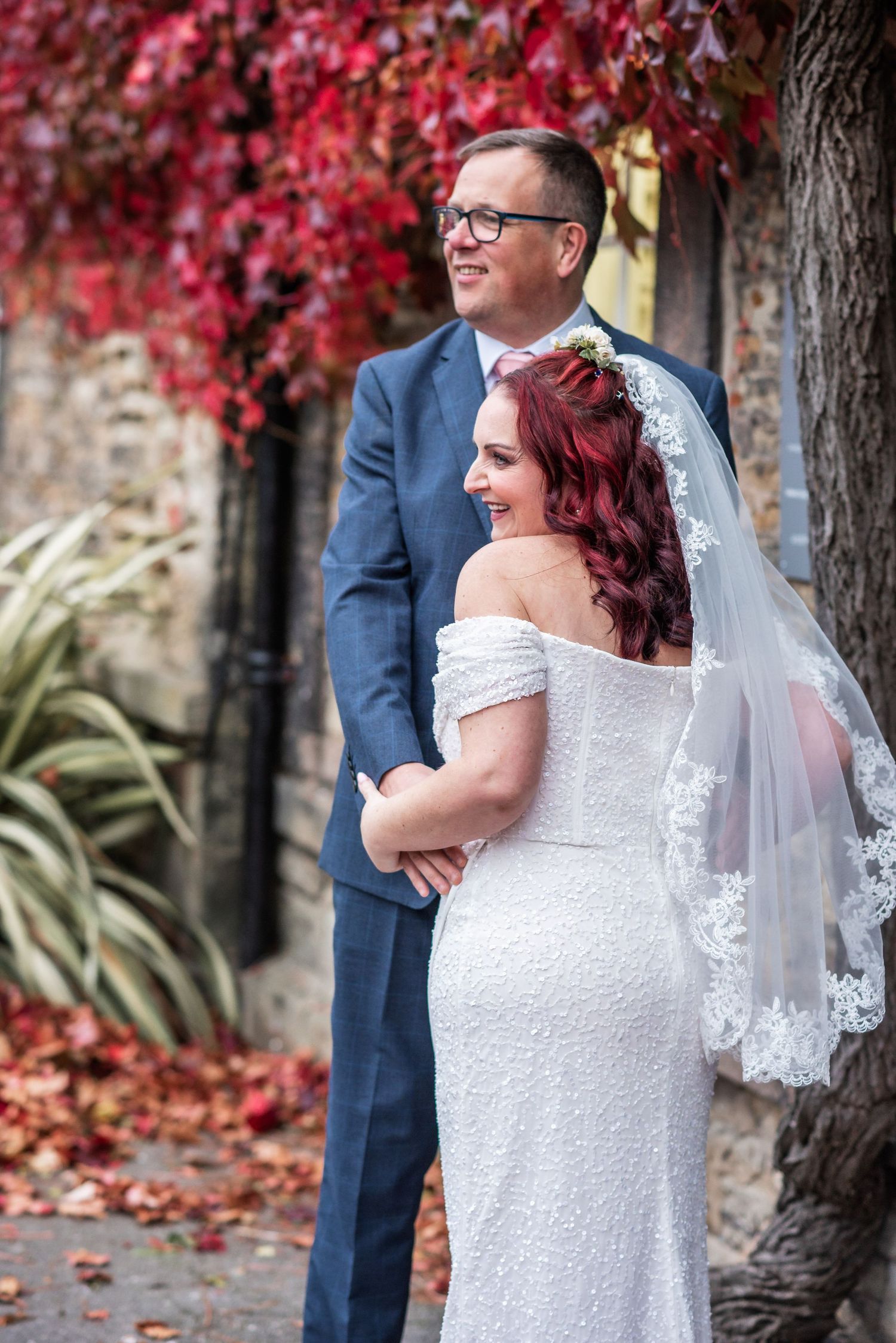 A newlywed couple shares a candid moment under bright red fall foliage and stone archway.
