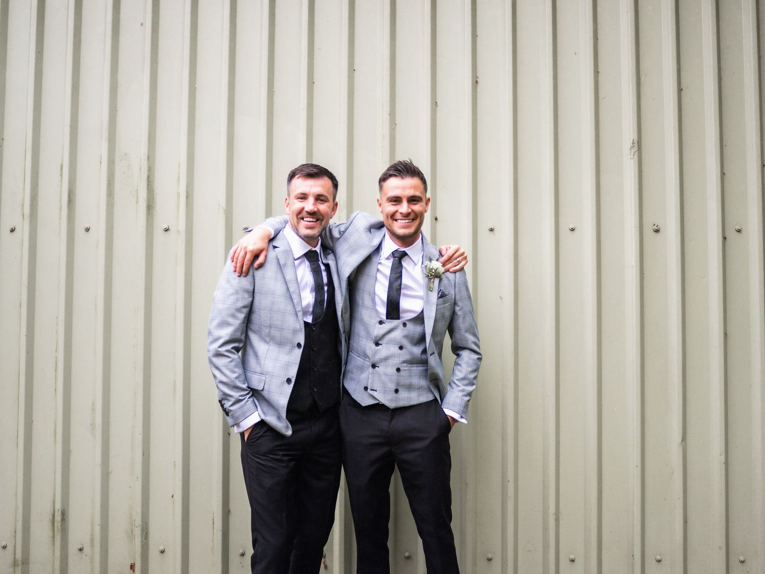 Two men in matching gray suits and navy ties pose against metal wall.