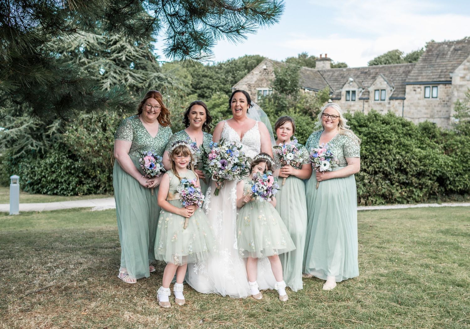 Bridal party in mint green dresses poses for a photo on a grassy lawn with stone buildings in background.