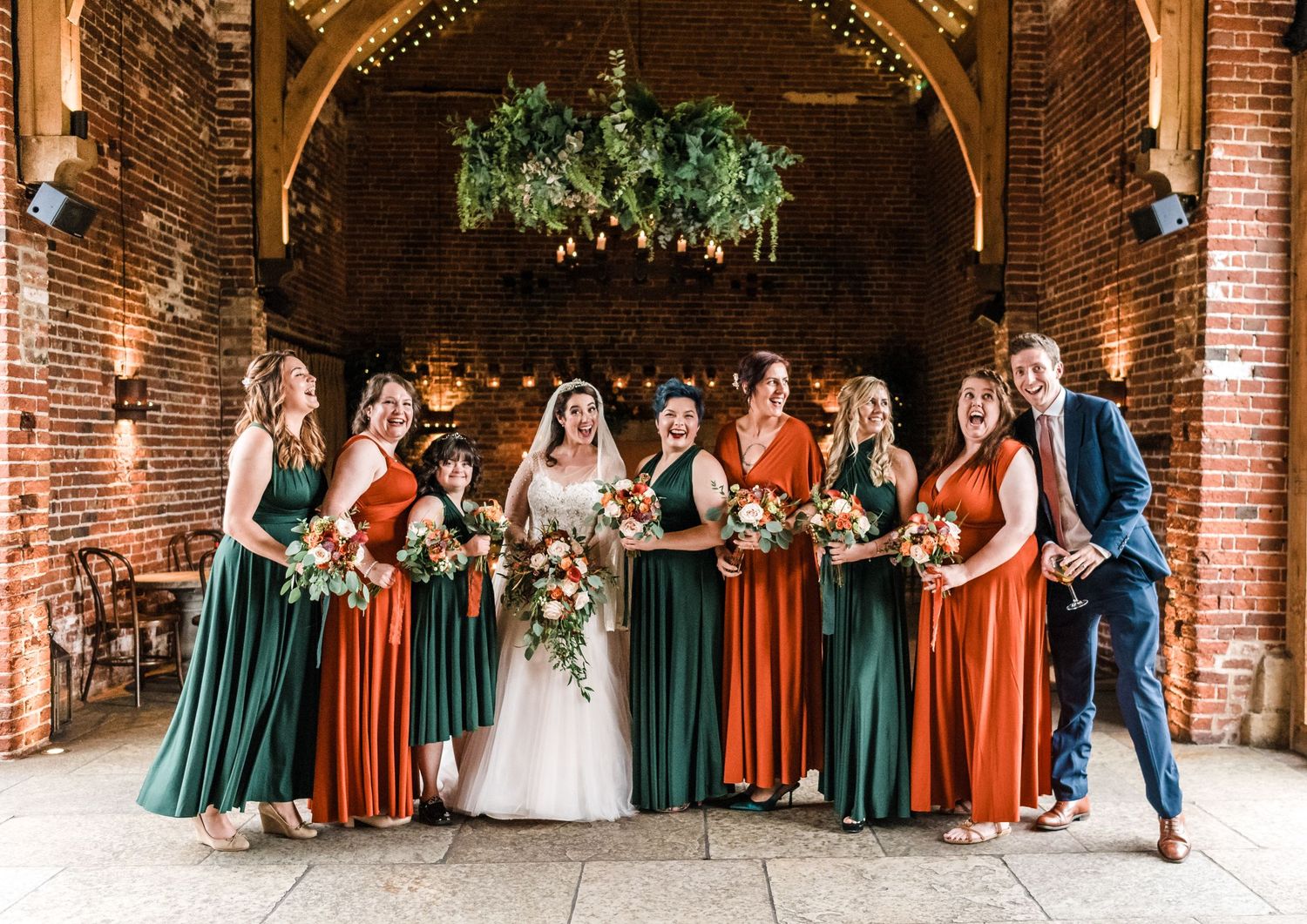 Wedding party poses under brick archway with bridesmaids in green and orange dresses.