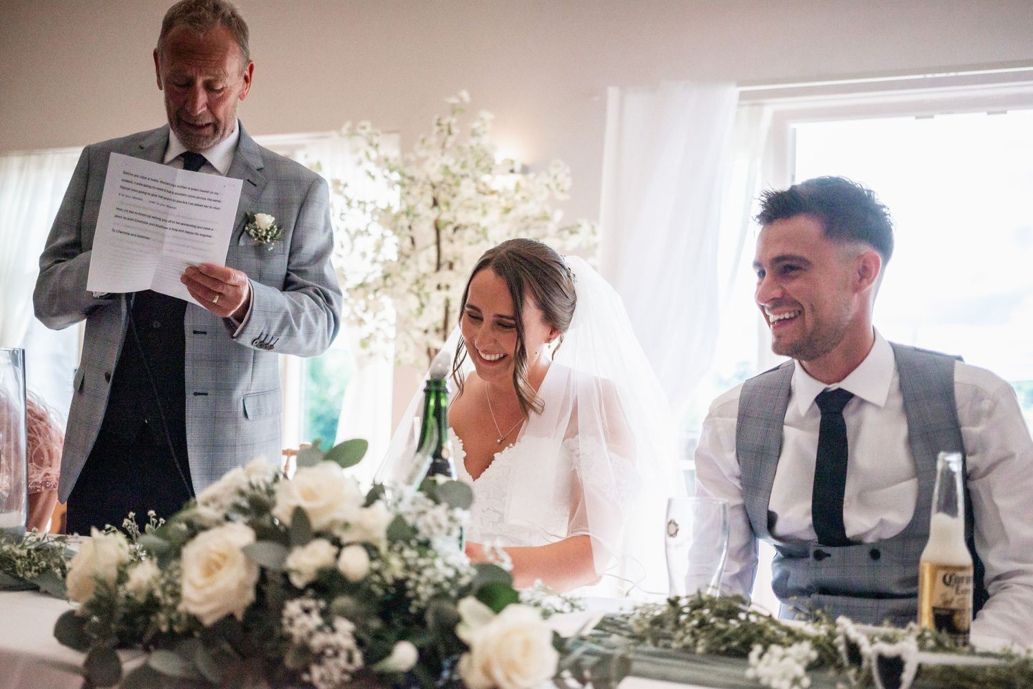 Wedding speeches and toasts during reception with floral centerpieces and wine bottles on table.