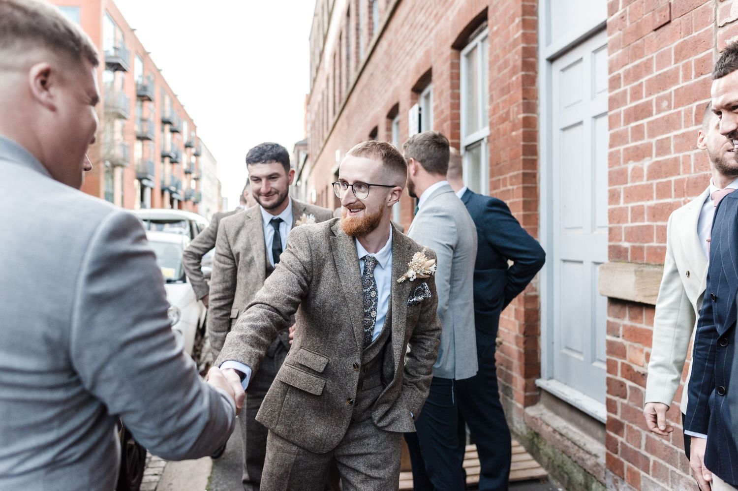 Wedding party members in suits greet each other outside a brick building.