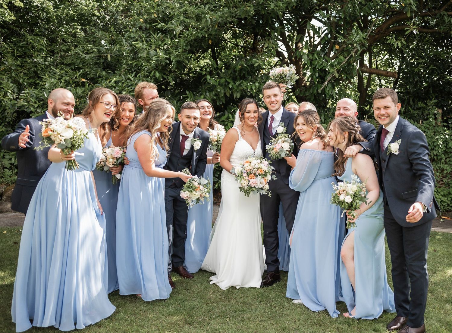 Wedding party in light blue attire poses together on a sunny garden lawn.