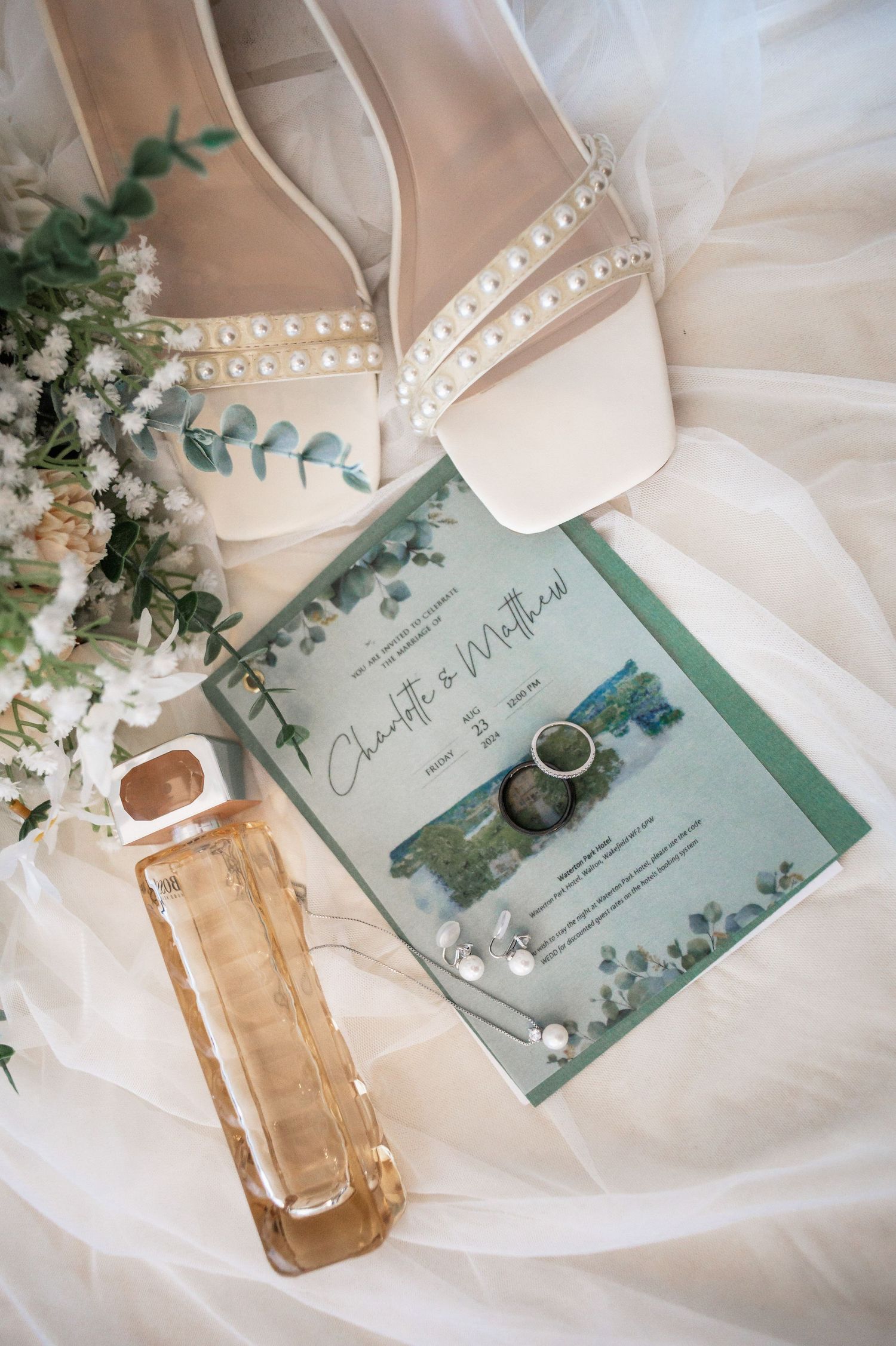 Wedding details flat lay with pearl shoes, invitation, perfume and eucalyptus on white fabric.