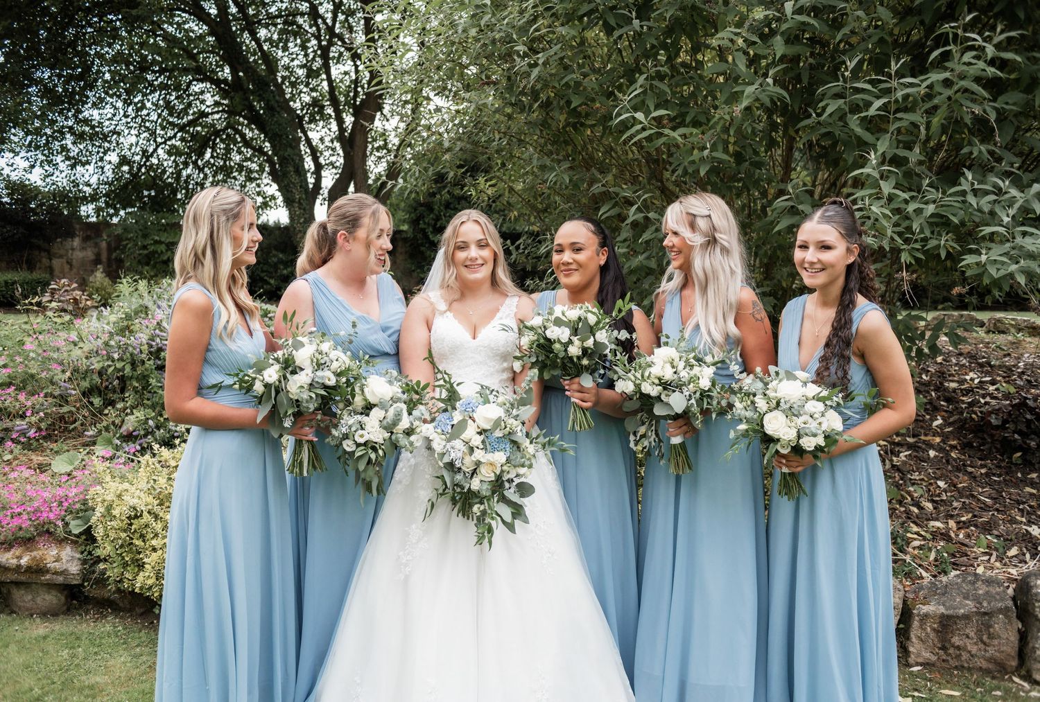 Wedding party in powder blue dresses holds white floral bouquets in garden setting.