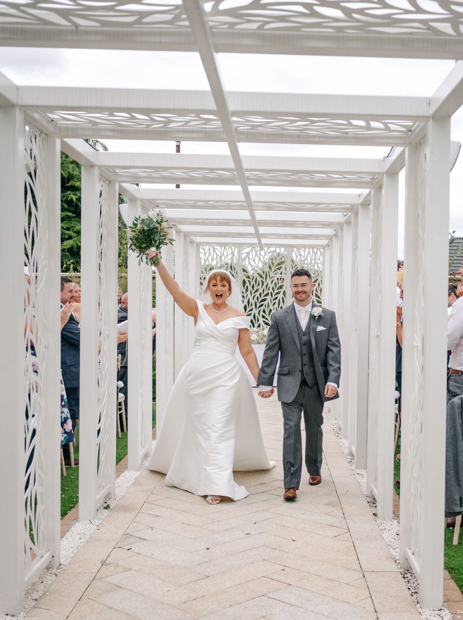 Newlyweds walking through an elegant white pergola with decorative leaf patterns.