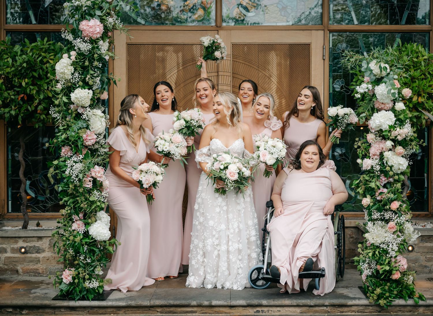 Wedding party in soft pink dresses seated on stone steps surrounded by elegant white floral arrangements.