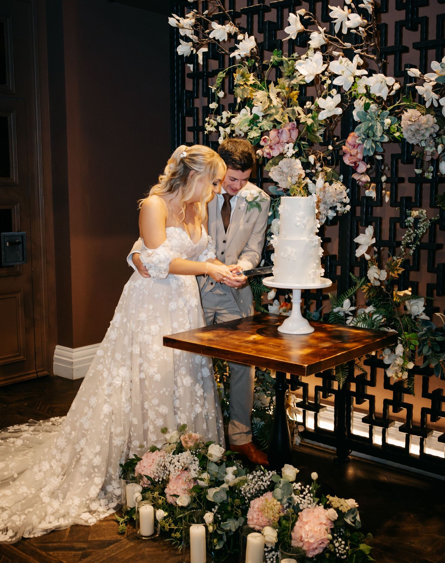 A wedding couple cuts their elegant white tiered cake surrounded by lush floral arrangements at their reception.