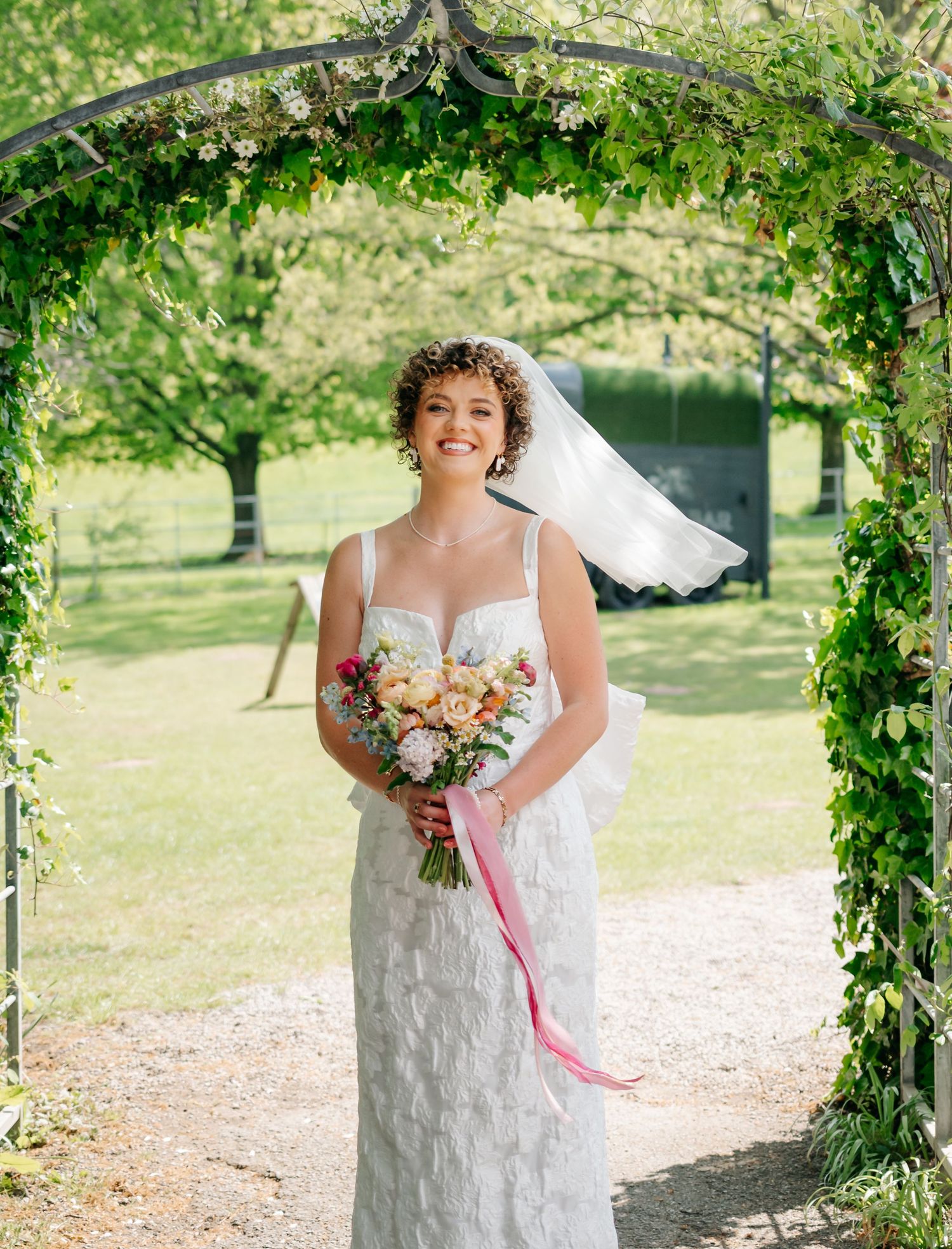Bride in white dress poses under a vine-covered garden arch on sunny day.