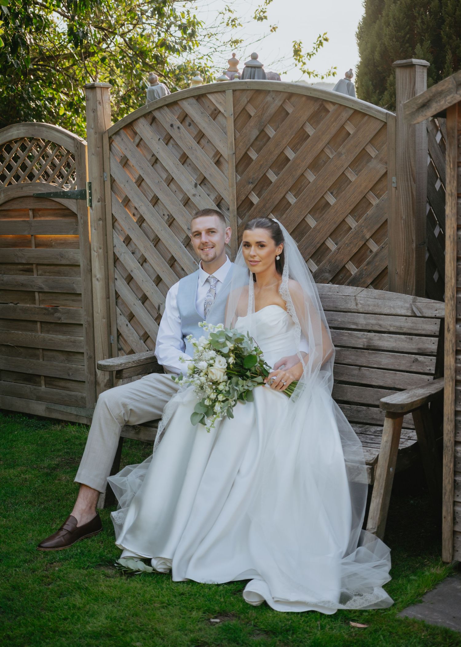 A couple poses on a wooden garden bench against a lattice backdrop with wedding florals.