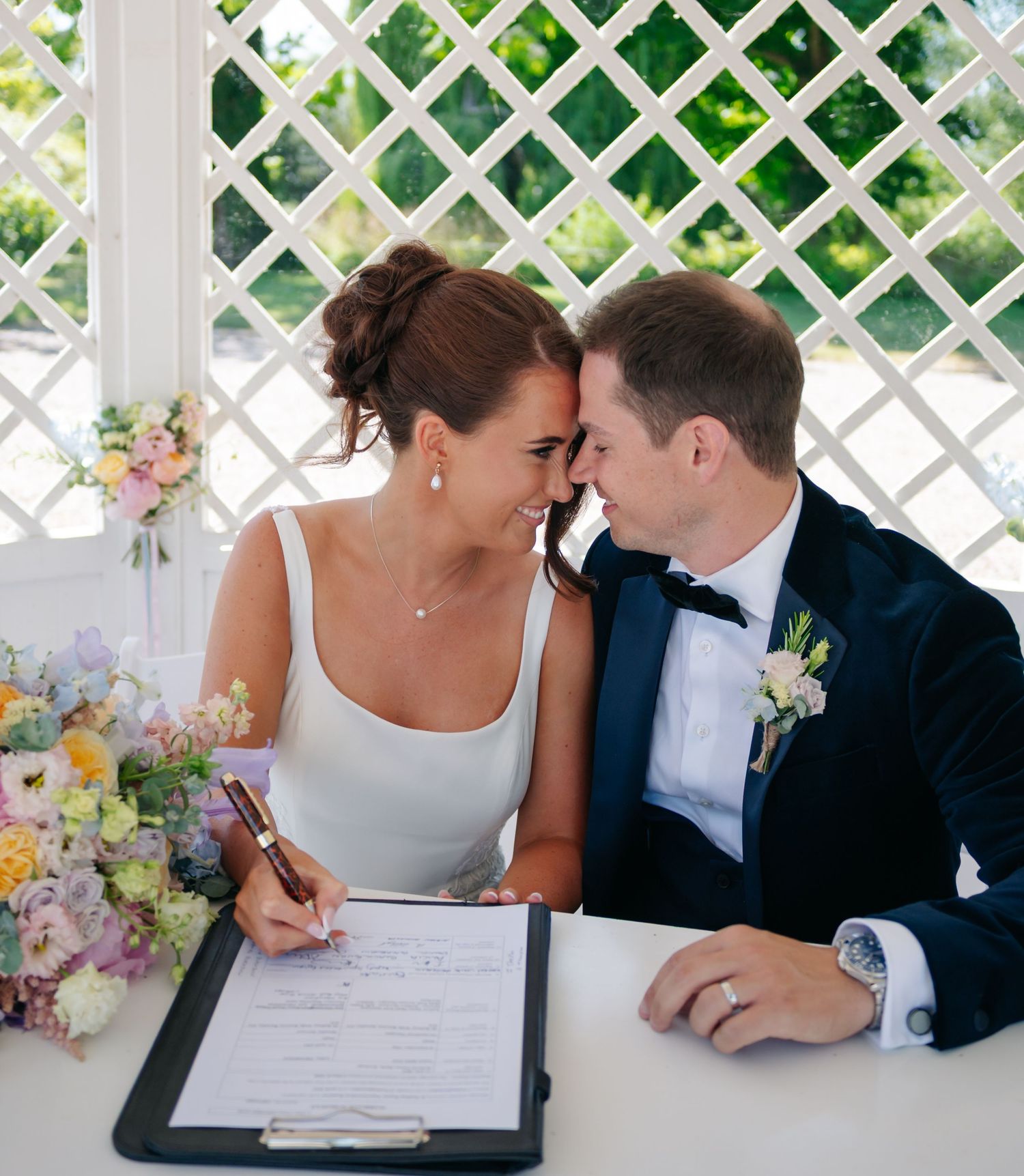 Newlyweds signing marriage documents together at a white lattice decorated ceremony space.