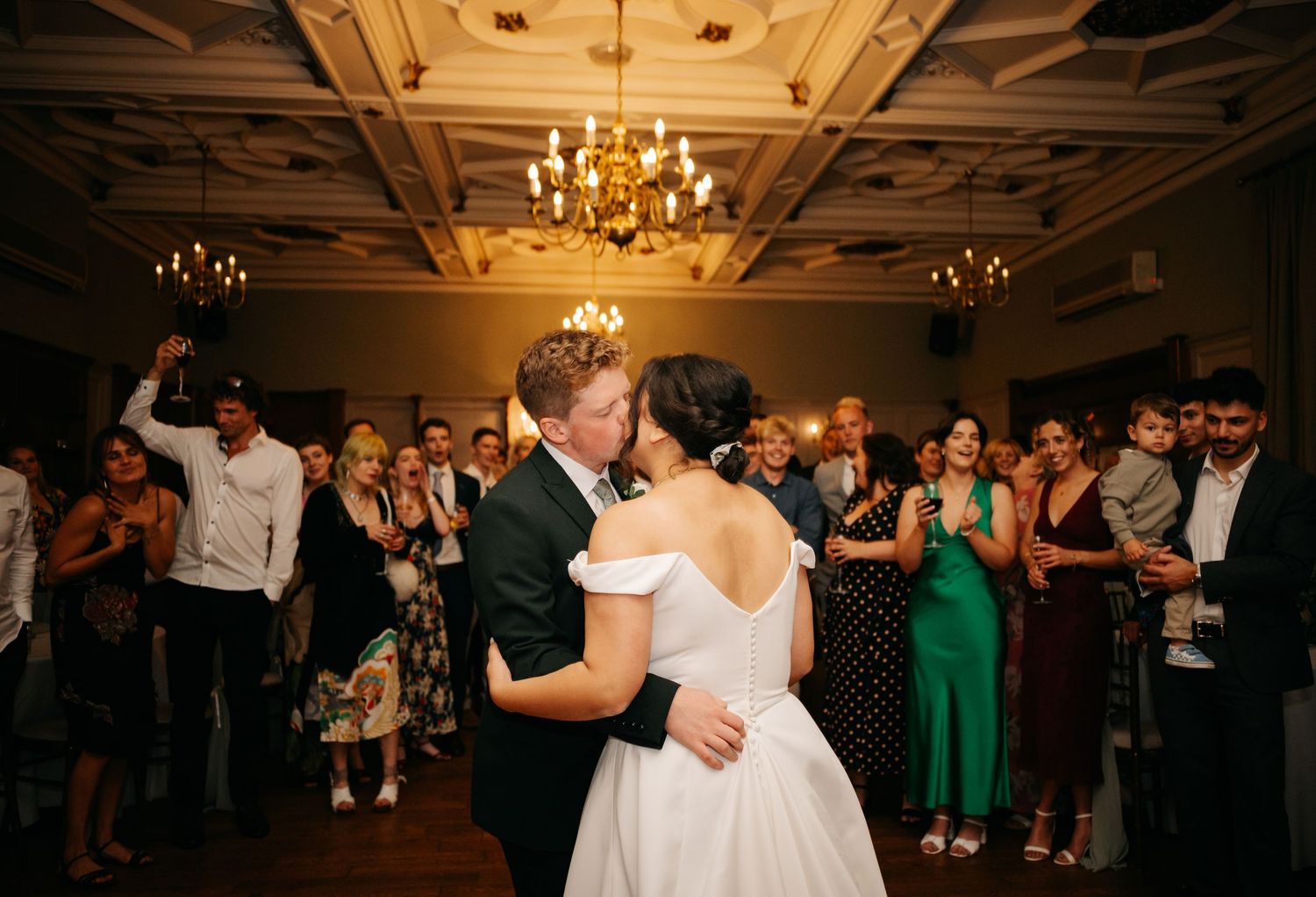 A couple shares their first dance at an elegant reception with chandeliers and guests watching.