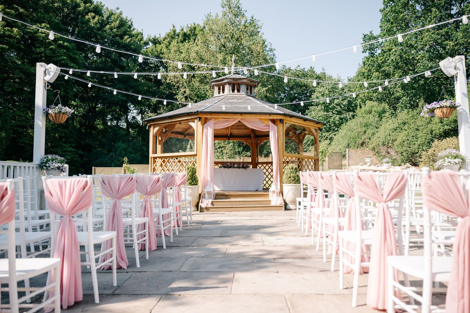 Outdoor wedding ceremony setup with white chairs adorned with pink sashes leading to an elegant wooden gazebo.