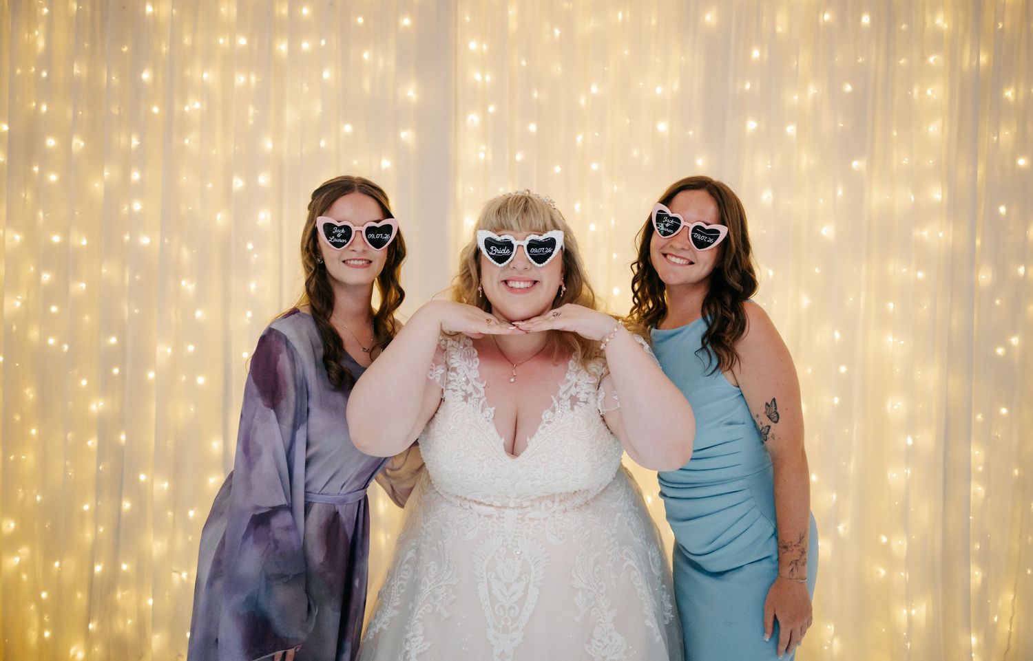 Bride in white gown poses with two bridesmaids wearing heart sunglasses in front of a glowing fairy light backdrop.