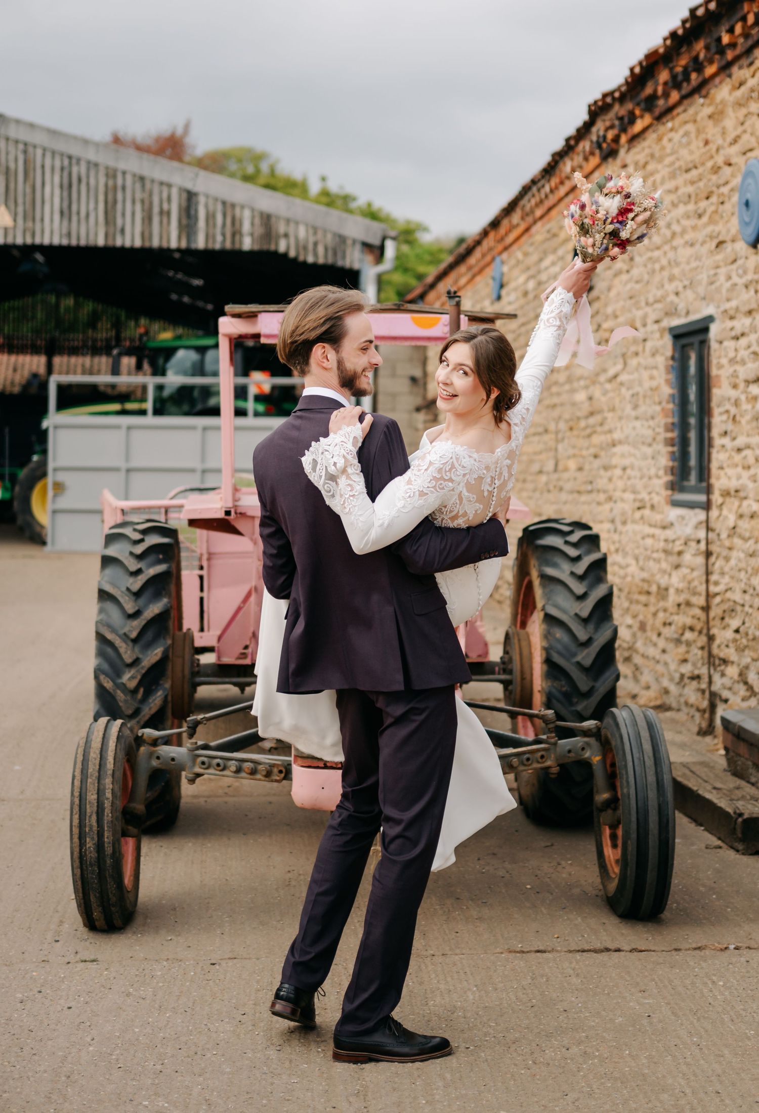 A couple shares romantic dance moves between vintage tractors on a rustic farm with brick buildings in background.
