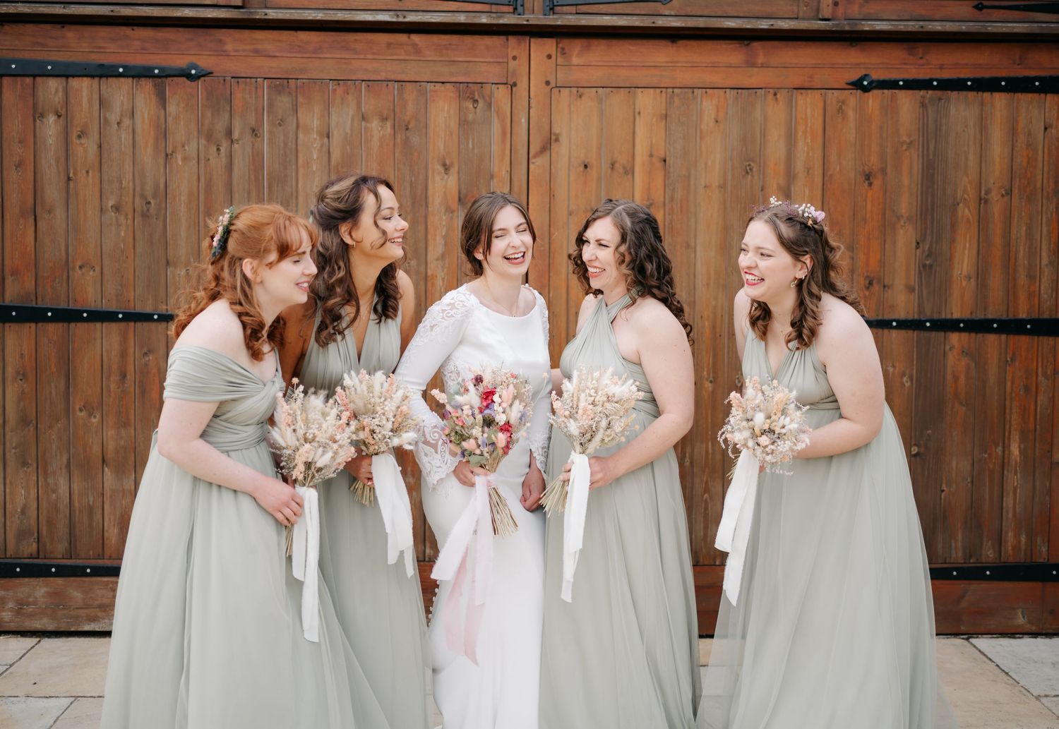 Bridesmaids in sage green dresses standing together against wooden barn backdrop.