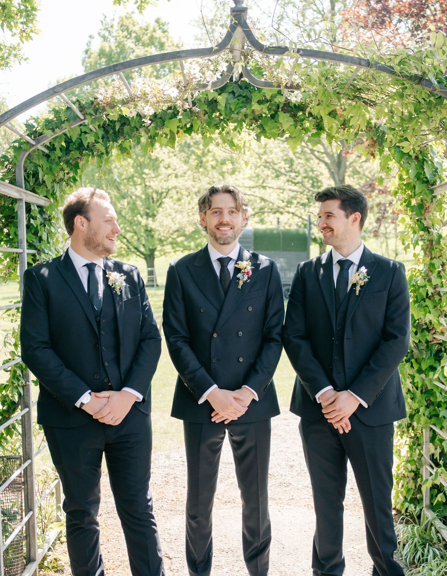 Groomsmen in black suits stand together under a garden arbor sharing candid moments.