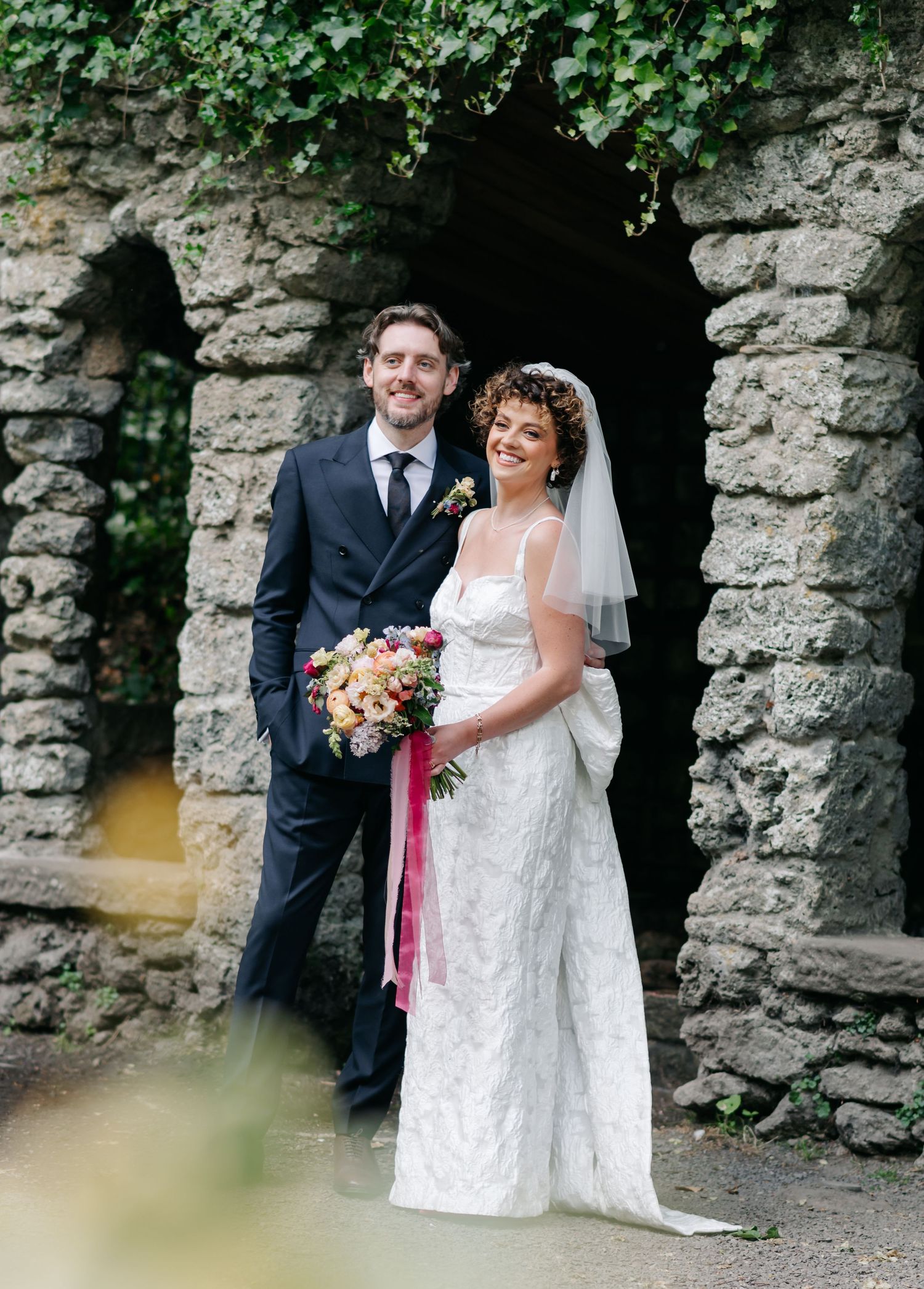 Wedding couple poses against a rustic stone wall backdrop with floral bouquet.