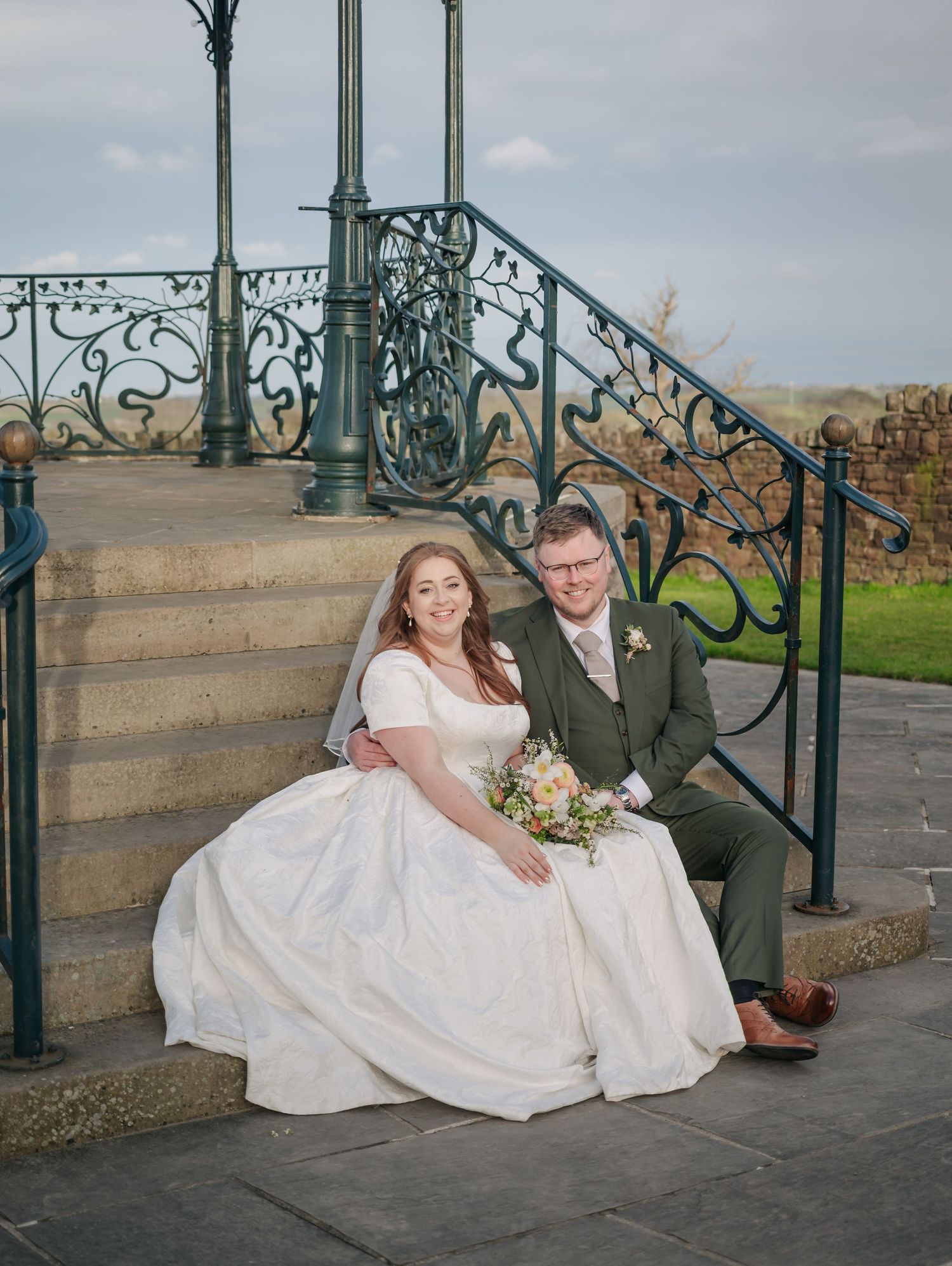 Newlyweds pose playfully on stone steps near ornate green metal railings during their wedding photoshoot.