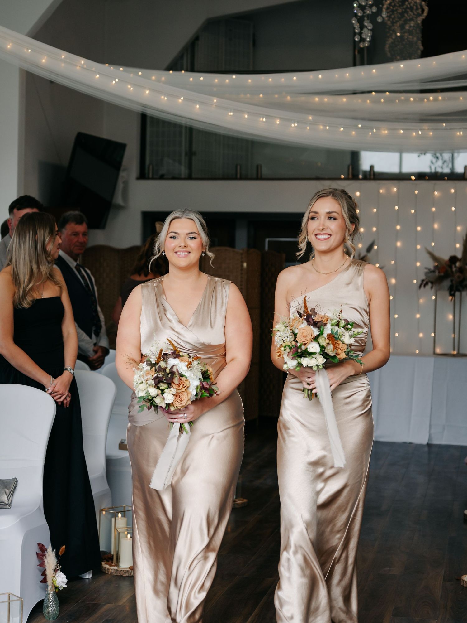 Bridesmaids in champagne silk dresses hold bouquets while laughing together during a wedding celebration with twinkle lights.