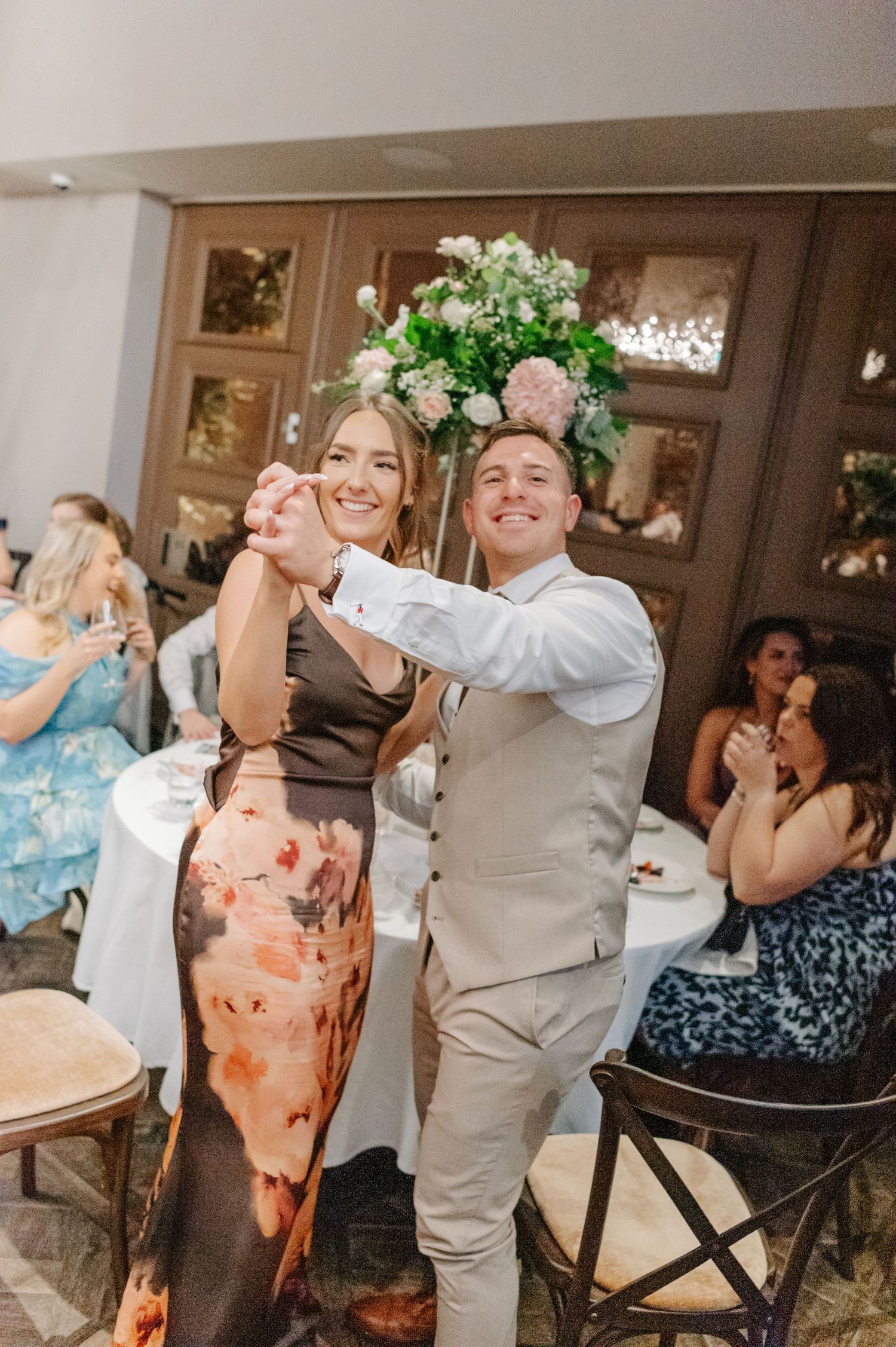 A couple dances joyfully at a wedding reception surrounded by floral arrangements and seated guests.