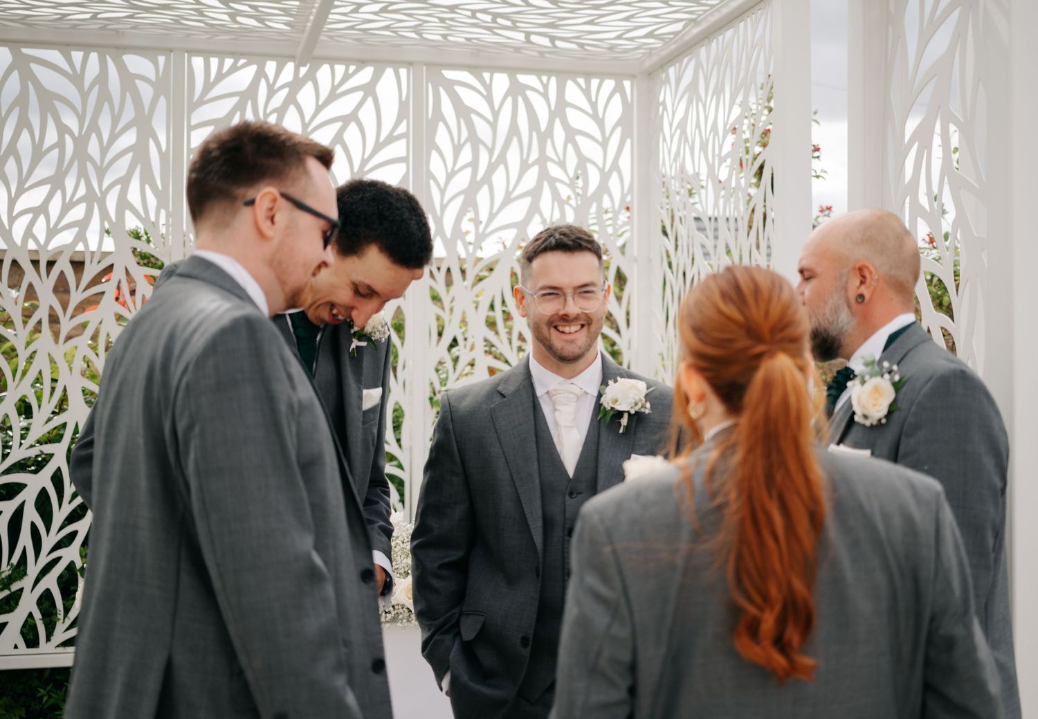 Wedding party gathered under a decorative white pergola with leaf cutout patterns.