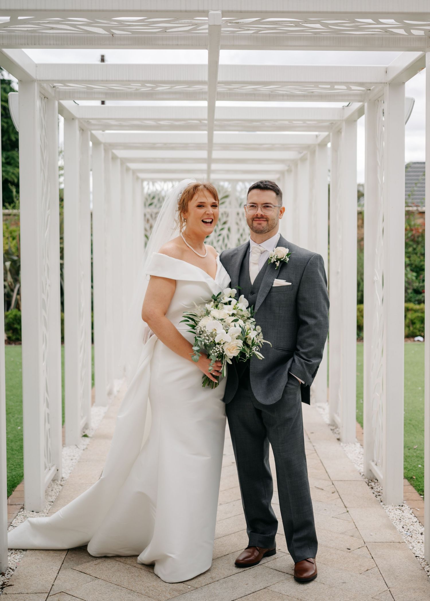 Wedding couple poses together under white pergola in garden setting with elegant floral bouquet and formal attire.