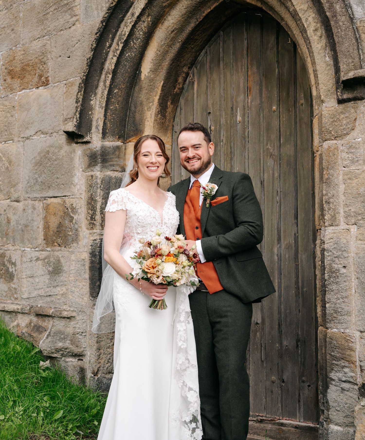 Bride in a lace wedding gown and groom in a green suit with orange waistcoat pose before an old stone church doorway.