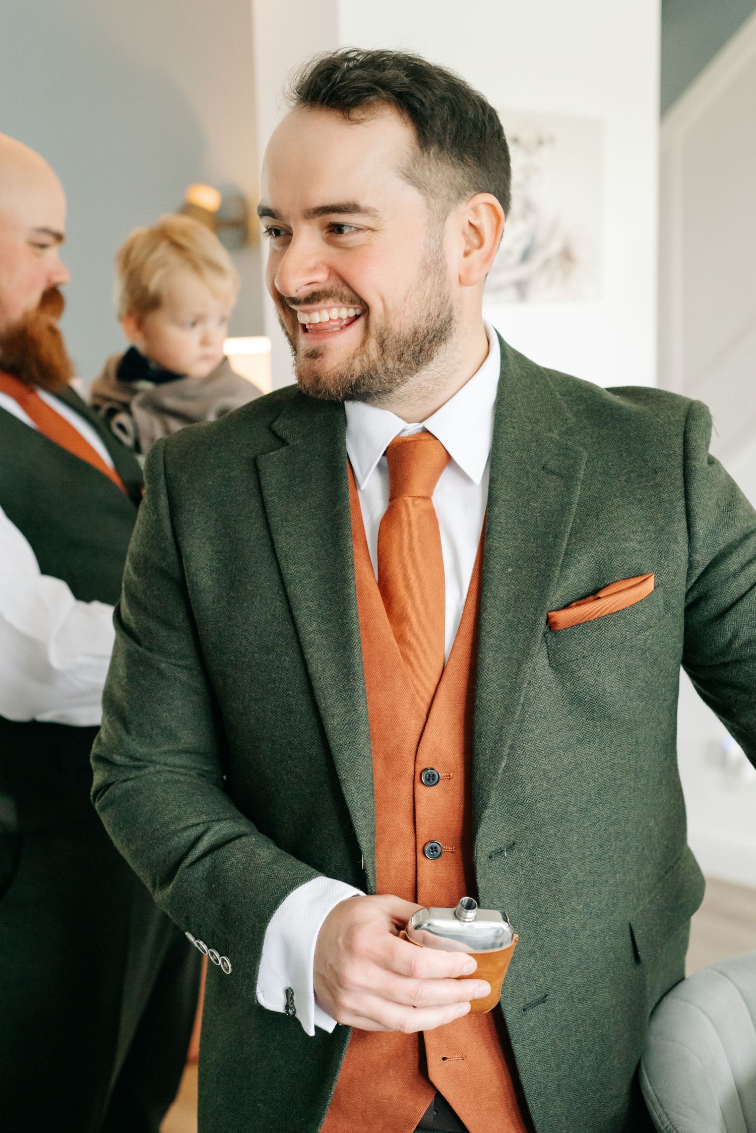 Smiling bearded groom in a green tweed jacket and orange vest holds a small flask while a child and another man stand behind him.