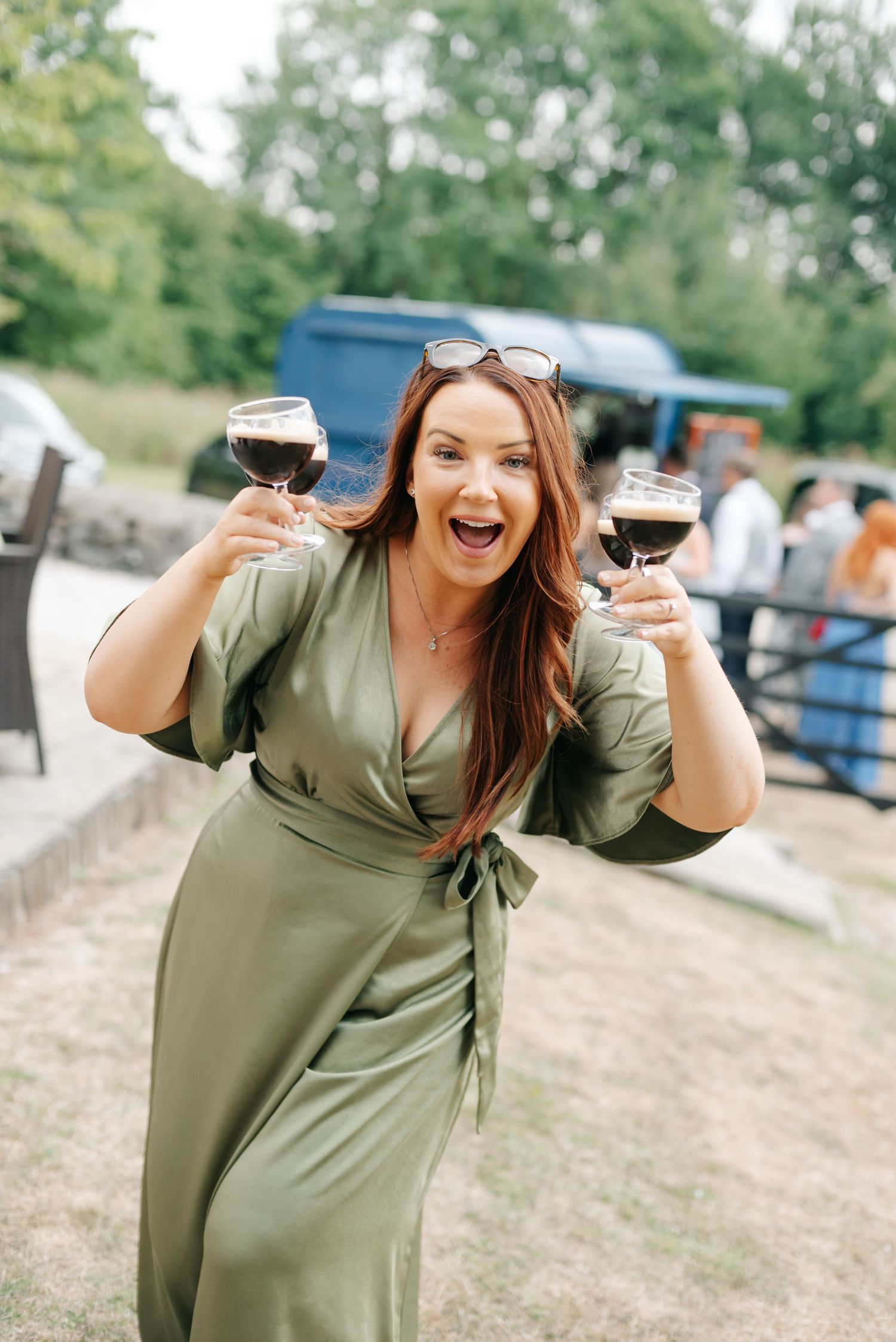 Excited woman in green wrap dress holding two glasses of red wine outdoors at a garden event.