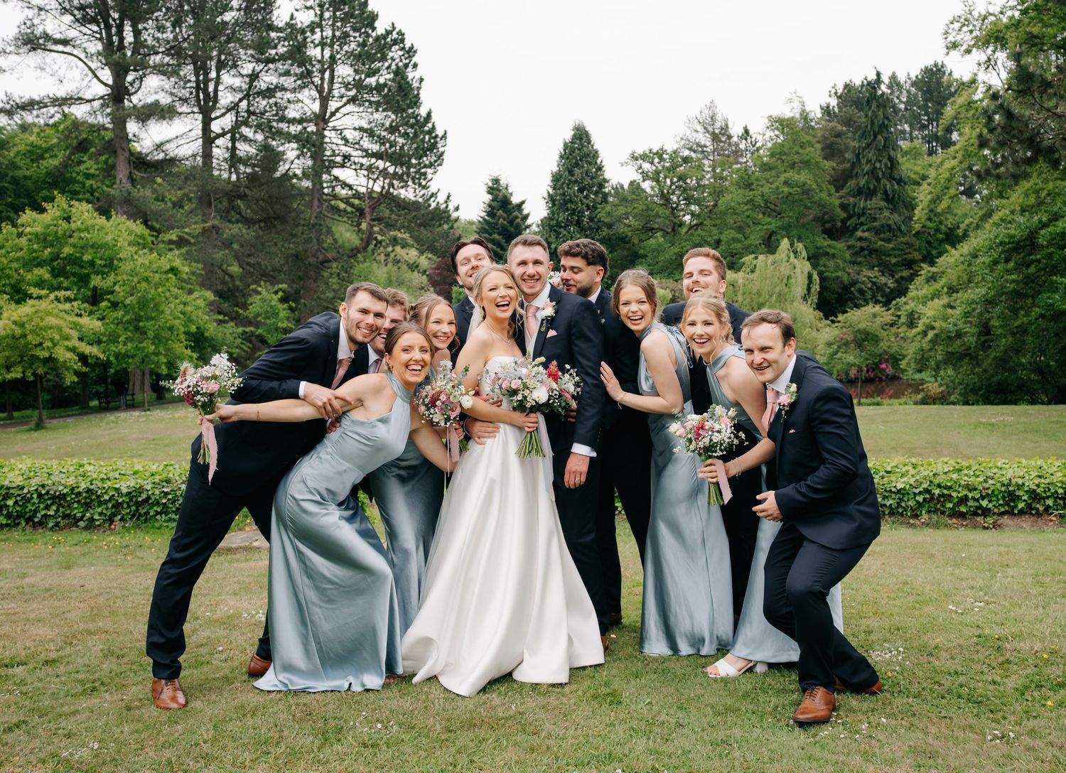 Wedding party poses playfully on a green lawn with bridesmaids in light blue dresses.