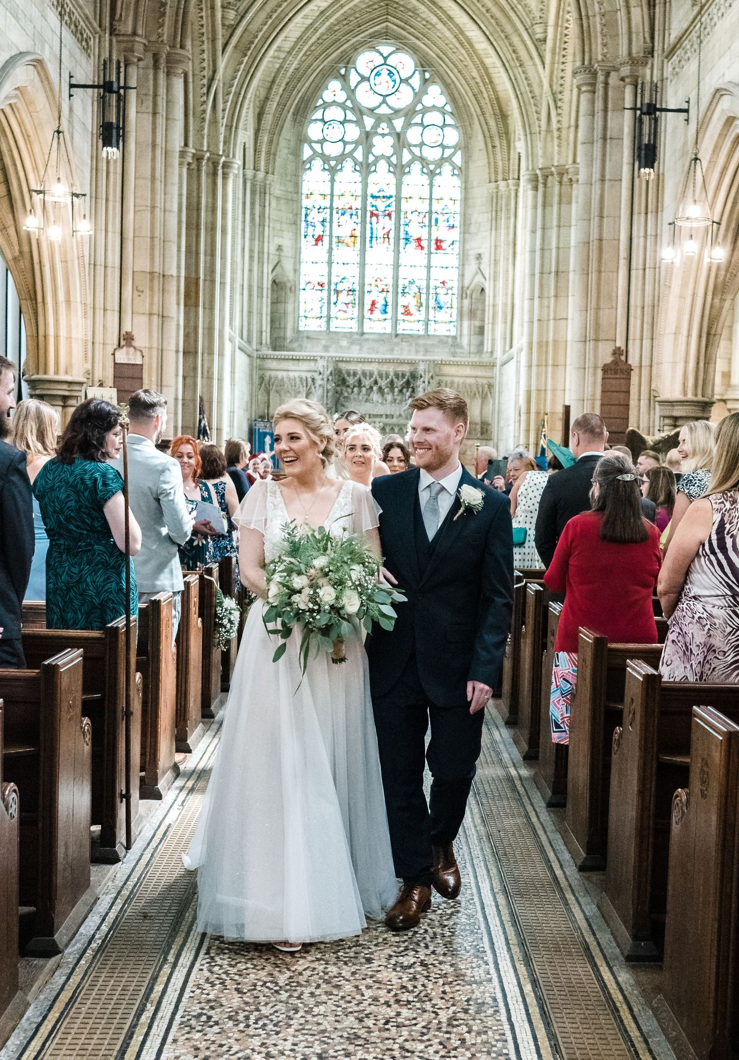 A couple walks down the aisle of a grand cathedral with stunning stained glass windows.