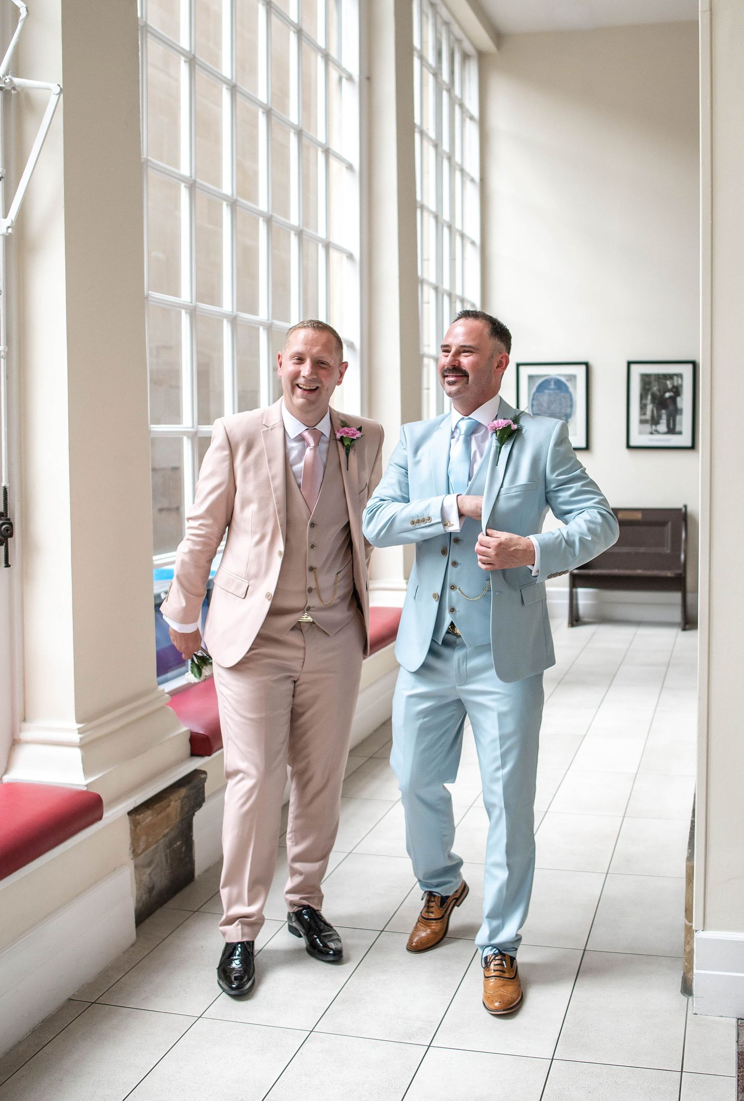 Two people walking and laughing together in pink and blue suits down a bright hallway.